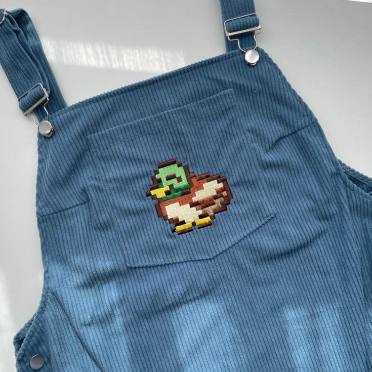 The Stardew Duck Overalls Dress by petimint is showcased, featuring a blue corduroy fabric with adjustable straps and metal buckles. The front pocket boasts a pixelated embroidered design of a duck, reminiscent of classic gaming graphics, in shades of green, brown, yellow, and white. The background is a light-colored surface.