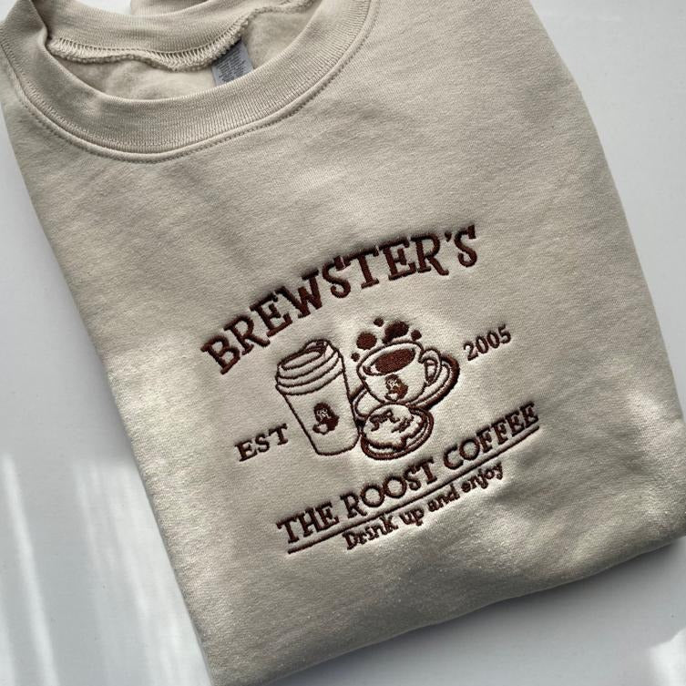 A folded beige sweatshirt, named Brewster's Crewneck by petimint, features embroidered text and images. It reads "BREWSTER'S EST 2005" above illustrations of a to-go coffee cup and a steaming coffee cup on a saucer. Below it says "THE ROOST COFFEE" and "Drink up and enjoy" in brown thread—perfect attire for every cozygamer's Nintendo Switch session.