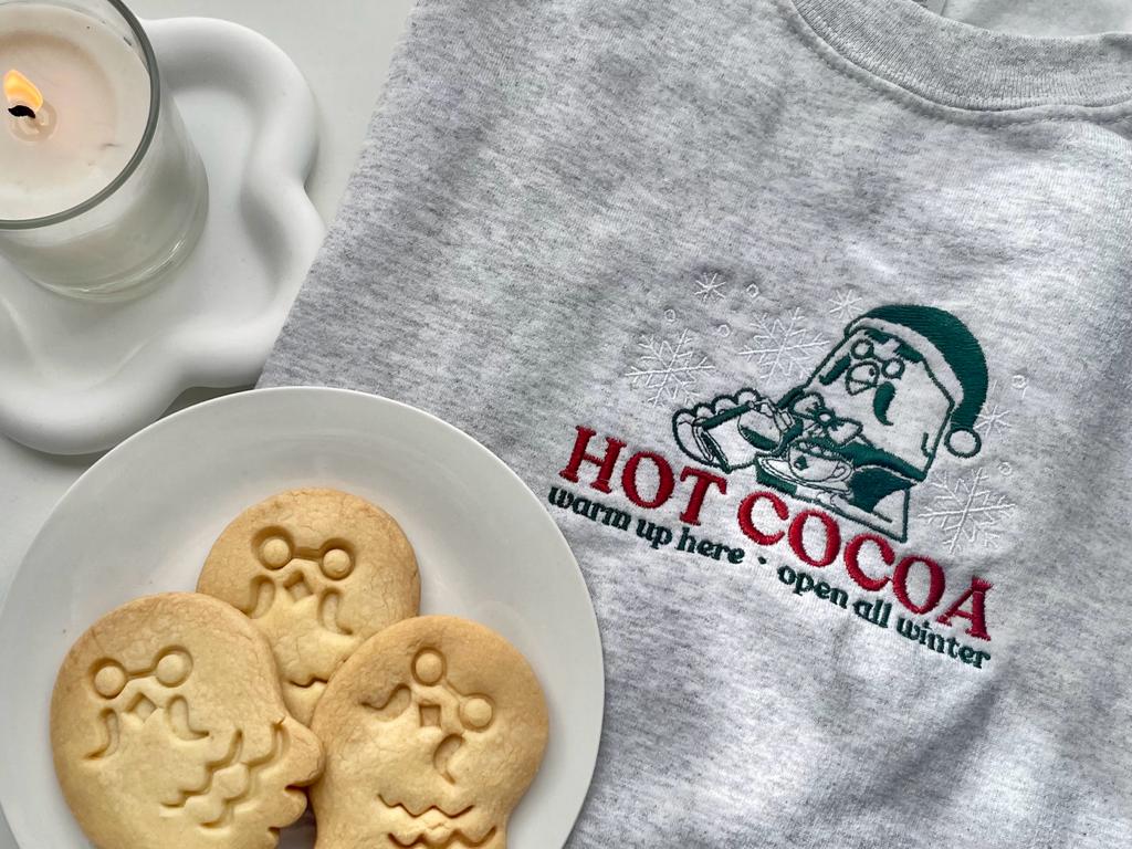 The petimint Hot Cocoa Sweatshirt, a light gray pullover featuring a festive design with an illustration of a character sipping hot cocoa and the text "HOT COCOA warm up here · open all winter," is displayed next to a lit candle, a white plate with three holiday-themed cookies, and a Nintendo Switch—perfect for the cozy gamer in your life.