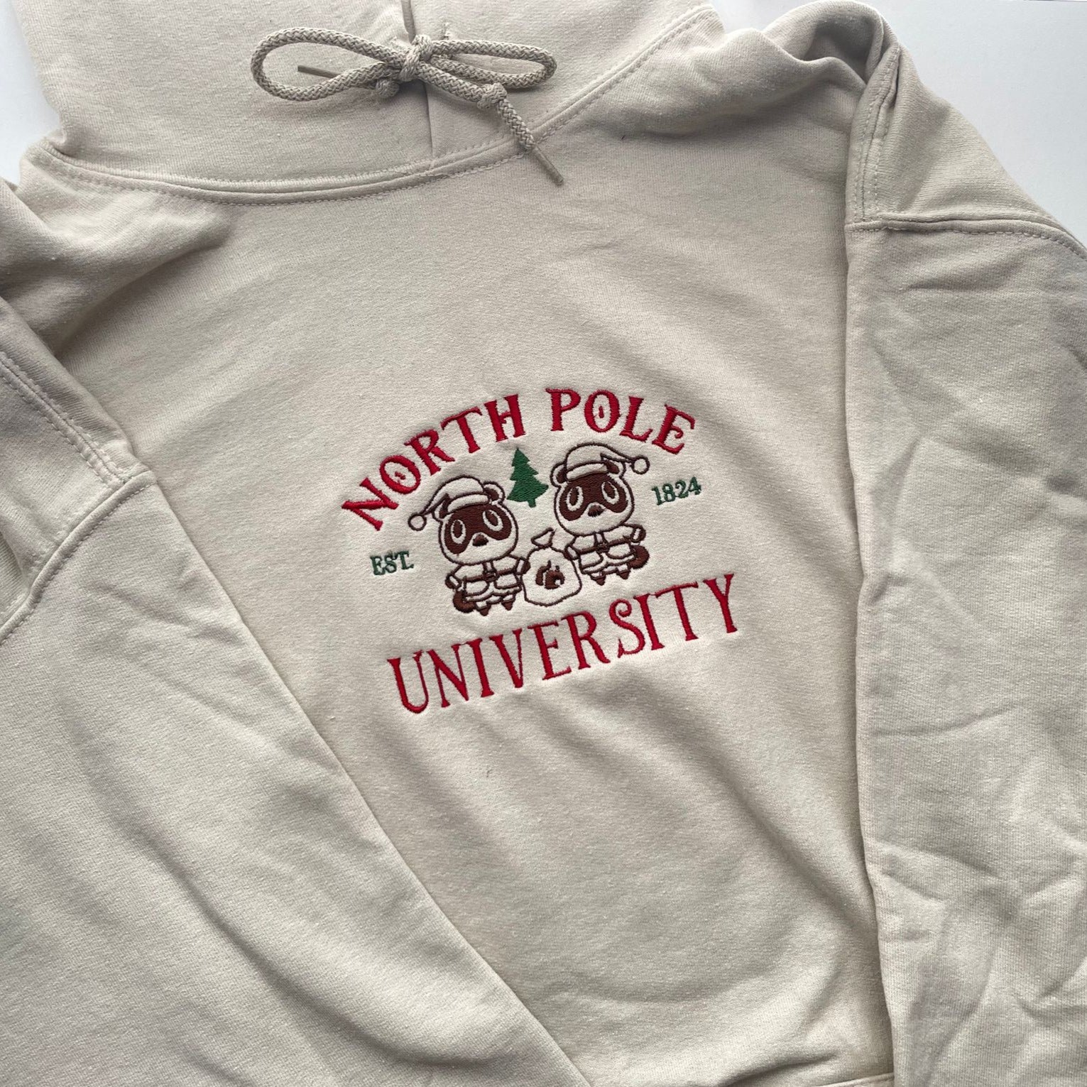 The North Pole University Hoodie by petimint is a light beige hoodie adorned with a festive design featuring two cartoon reindeer dressed in academic attire. The text "North Pole University" is written in red, with "EST. 1824" in green. Perfect for cozygamer apparel, it's ideal for lounging and playing Nintendo Switch. Spread out on a surface.