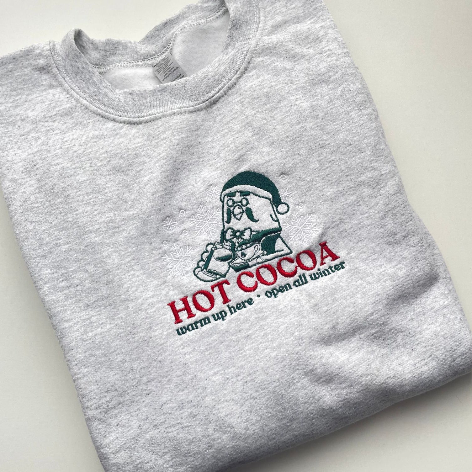 The petimint Hot Cocoa Sweatshirt, in ash grey, features a festive design with a cartoon character wearing a winter hat and glasses, holding a mug. The text "HOT COCOA warm up here - open all winter" is embroidered in red and green, making it the perfect piece for the #cozygamer.