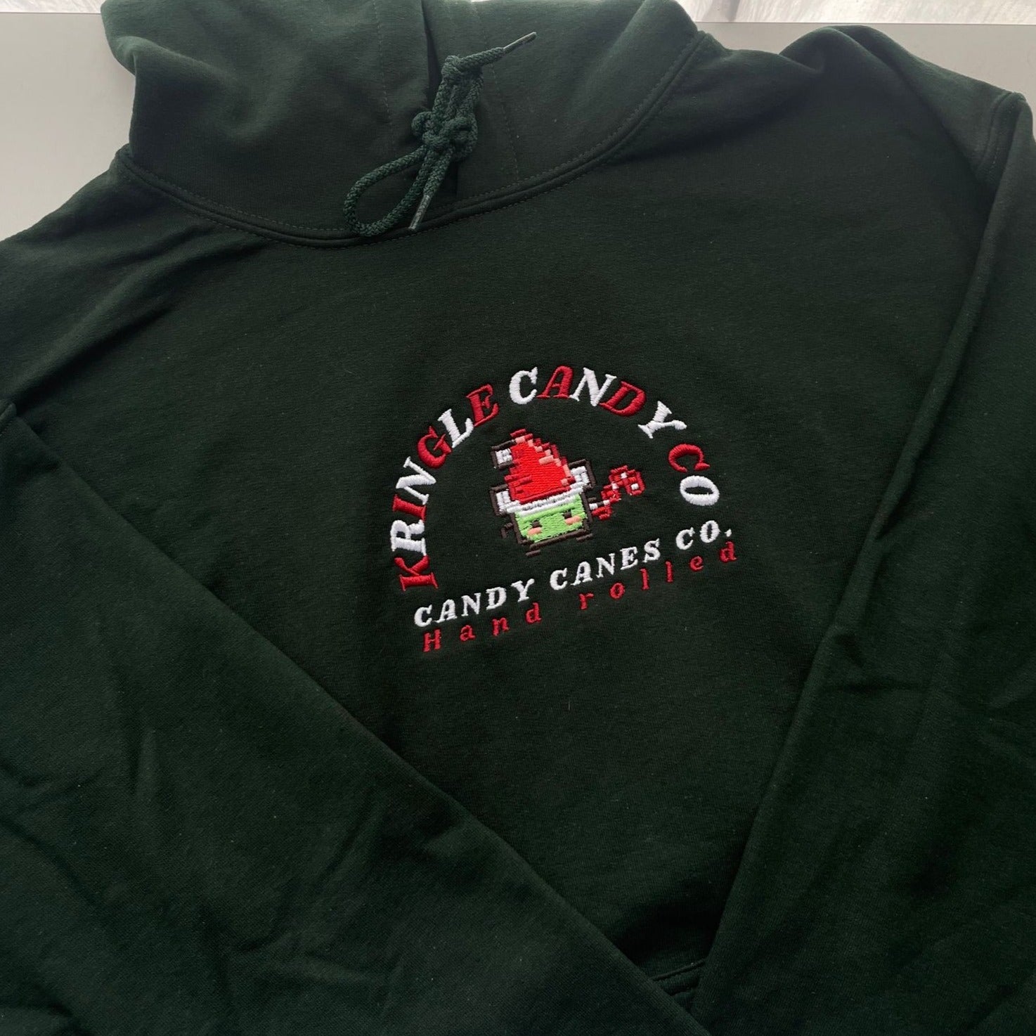 The petimint Candy Canes Junimo Hoodie is a dark green hoodie featuring a colorful embroidered design on the front. At the center of the design is a red candy cane being made, surrounded by "Kringle Candy Co." and "Candy Canes Co., Hand Rolled" in red and white embroidery. Ideal for gamers, this cozy piece of apparel seamlessly blends festive fun with casual comfort.