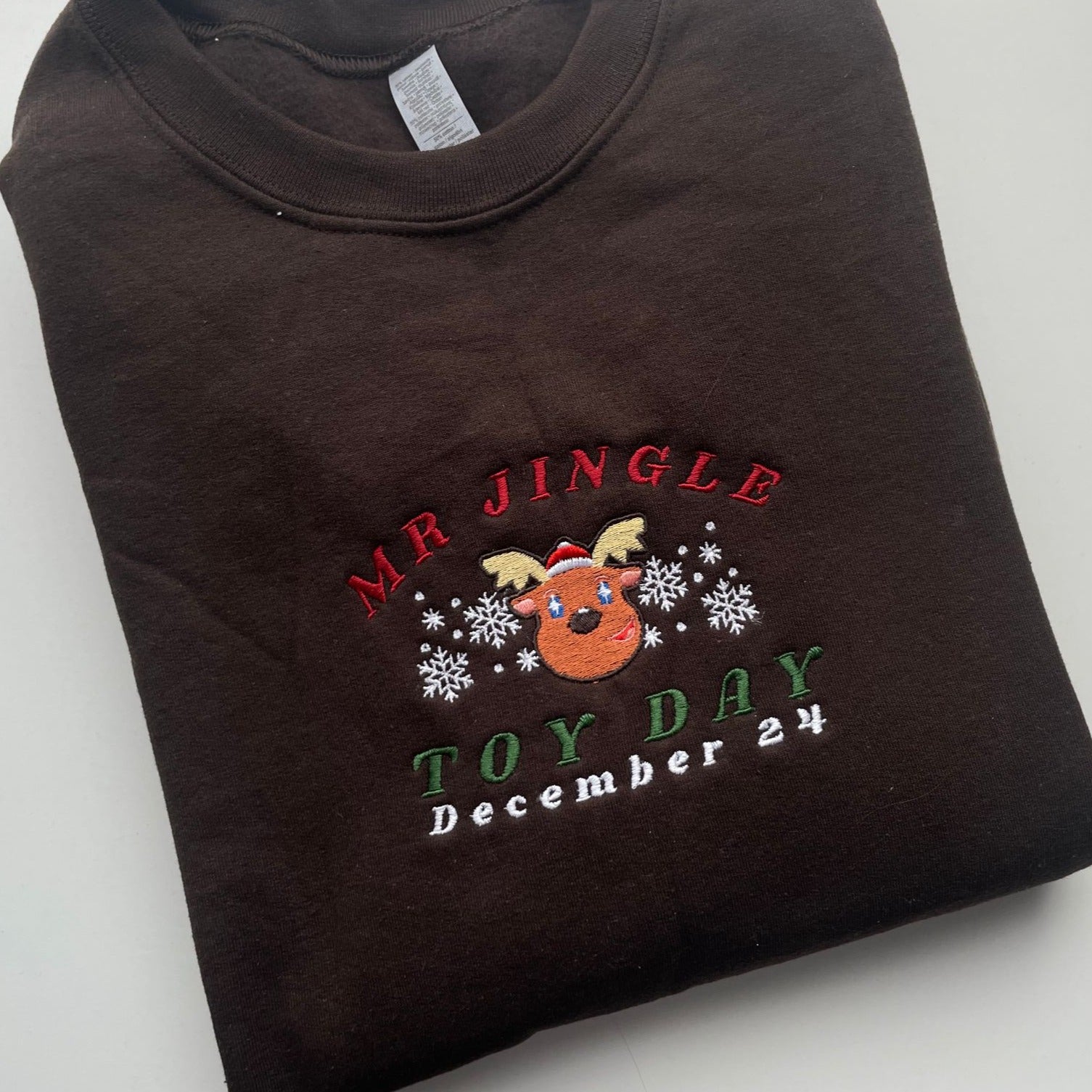 A folded dark brown "Mrs Jingle Sweatshirt Christmas Apparel" by petimint features an embroidered design of a smiling reindeer head with snowflakes around it. The design includes the words "Mr. Jingle" in red and "Toy Day December 24" in green and white, making it perfect for gaming sessions on your Nintendo Switch or any cozy day.