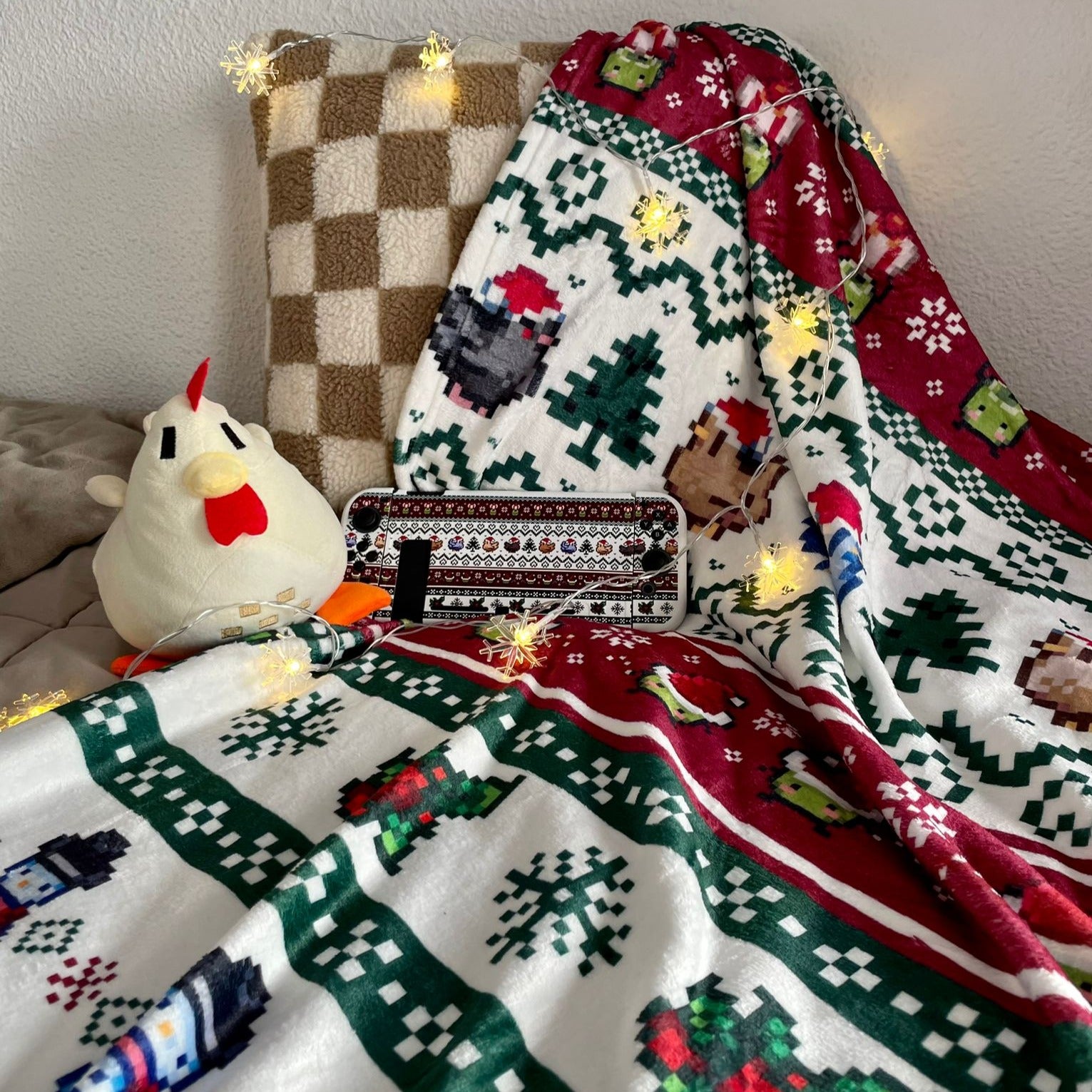 A cozy gaming scene with a petimint Stardew Christmas Blanket, featuring festive red, green, and white colors, draped over a beige checkered pillow. A plush chicken toy and a Nintendo Switch adorned with a festive cover are placed on the blanket, surrounded by warm, glowing fairy lights.