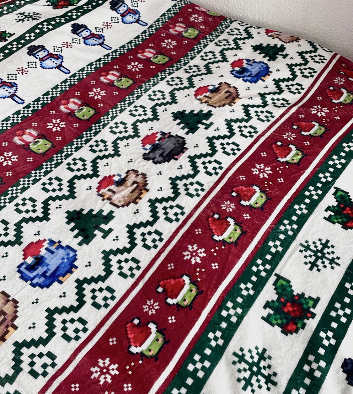 The Stardew Christmas Blanket by petimint is a festive fabric with a Christmas-themed design featuring horizontal rows of pixelated characters, including snowmen, owls in Santa hats, Christmas trees, and snowflakes in colors of red, green, white, blue, and brown—perfect for cozygamer apparel.