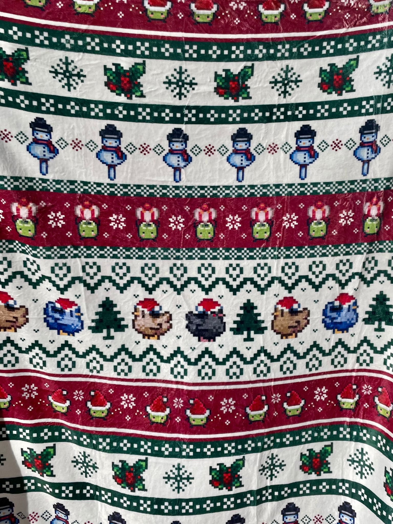 The Stardew Christmas Blanket by petimint boasts a festive pattern with horizontal rows showcasing snowmen, holly leaves, green Christmas trees, and bears wearing Santa hats. The vibrant colors include green, white, red, and blue arranged in an alternating sequence—perfect for a cozy holiday gaming night with your Nintendo Switch.