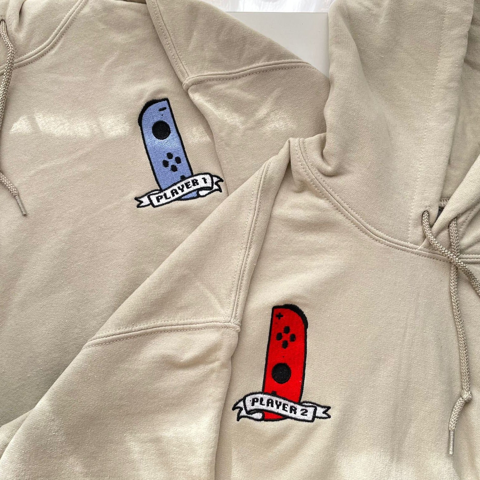 Displayed are two beige hoodies from the petimint collection, part of the "Pack 2 Hoodie Player 1 & 2 Gamer Apparel" set. Each hoodie features an embroidered design of a game controller on the chest—one in blue labeled "Player 1" and the other in red labeled "Player 2." Perfect for any gamer, this gaming apparel showcases controllers with similar shapes and button layouts.