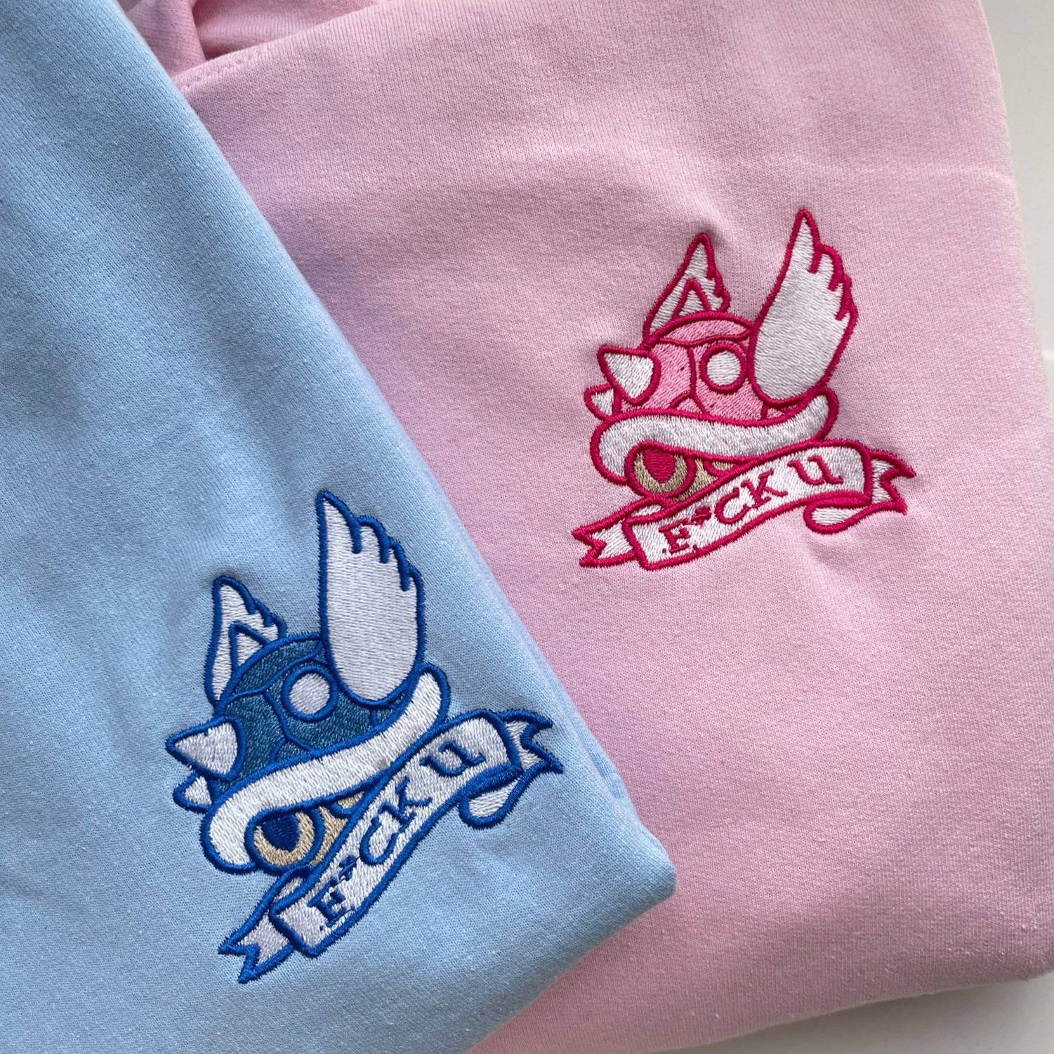 Two sweatshirts are pictured: one light blue and one Pink F*ck U Sweatshirt Gamer Apparel from petimint. Both feature an embroidered emblem of a crowned turtle shell with wings, along with a banner below it that reads "F*CK U" in stylized red and white lettering. Perfect apparel for the cozy gamer who loves their Nintendo Switch sessions.