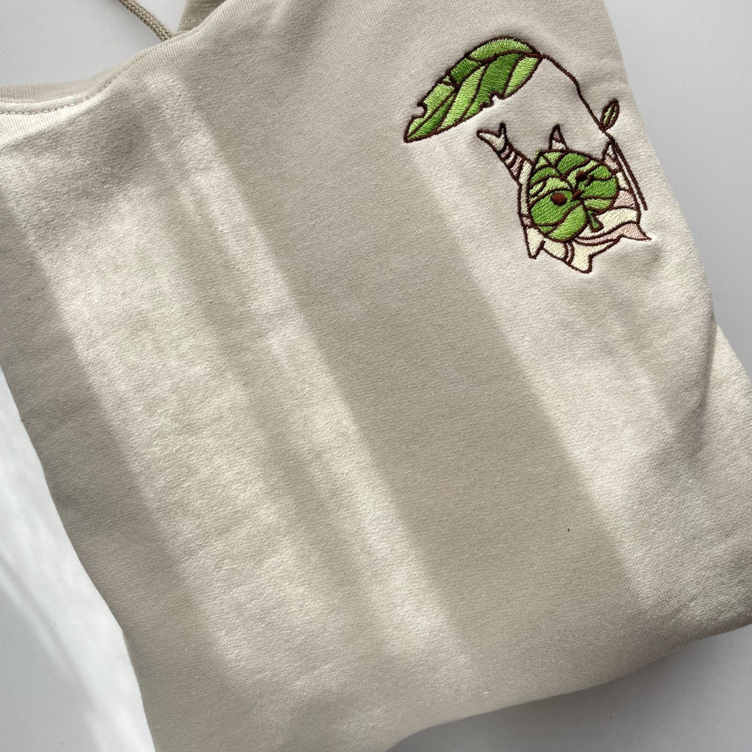 A beige Korok Hoodie by petimint, featuring an embroidered design of a small green leaf character hanging from a larger green leaf, is ideal for gaming enthusiasts. Positioned on a white surface, the stylish hoodie is bathed in natural light that casts subtle shadows across it.