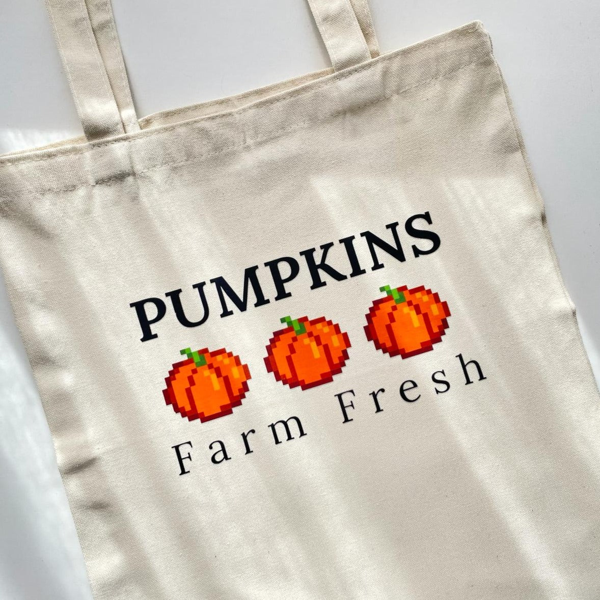 The Pumpkins Tote Bag Halloween by petimint is shown laid flat on a white surface, featuring the text "PUMPKINS Farm Fresh" accompanied by three pixelated pumpkin illustrations. This beige tote is crafted from durable polypropylene non-woven fabric and is heat-sealed for added durability.