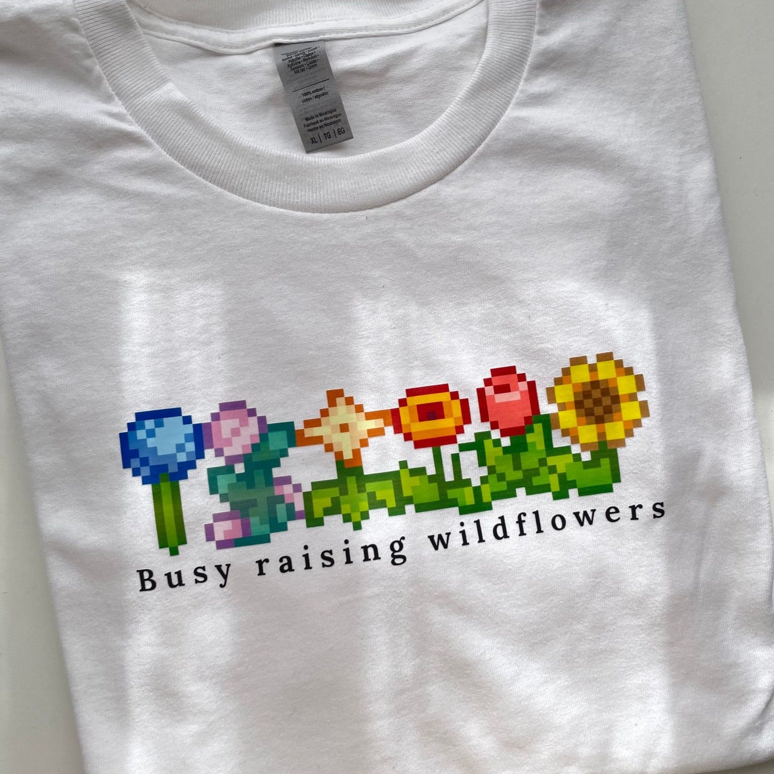 The Wildflowers Stardew T-Shirt by petimint showcases a row of pixelated flowers in various colors on the front, evoking the enchanting aesthetic of Stardew Valley. Beneath the flowers, the text "Busy raising wildflowers" adds a whimsical touch to this white tee, making it a delightful addition to your wardrobe.