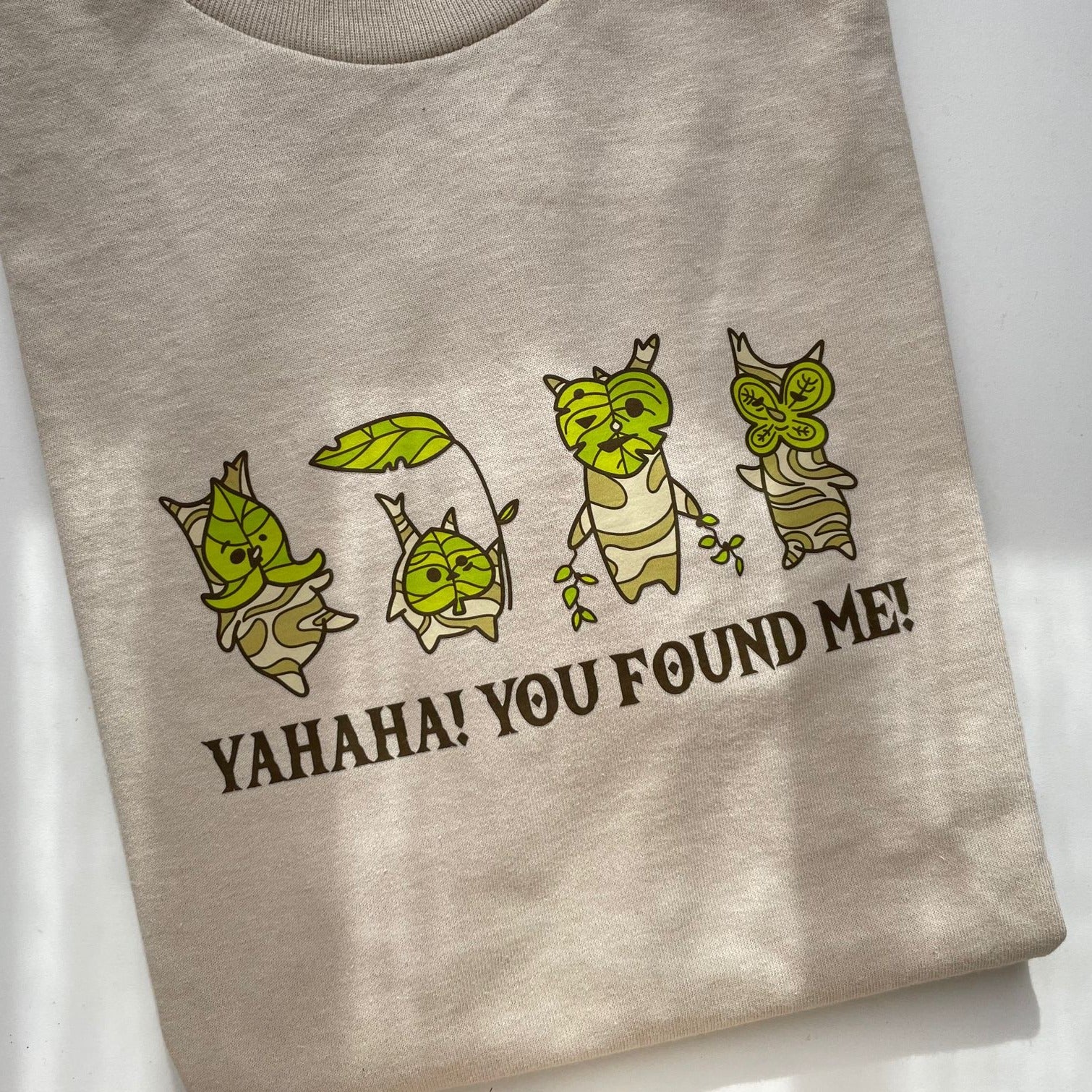 The Korok Yahaha! T-Shirt by petimint is a beige top adorned with four whimsical green and brown characters showcasing leafy details, each in a unique pose. Below them, the bold capital text reads "Yahaha! You Found Me!" Perfect for any gamer, the fabric feels soft and the design exudes playfulness.