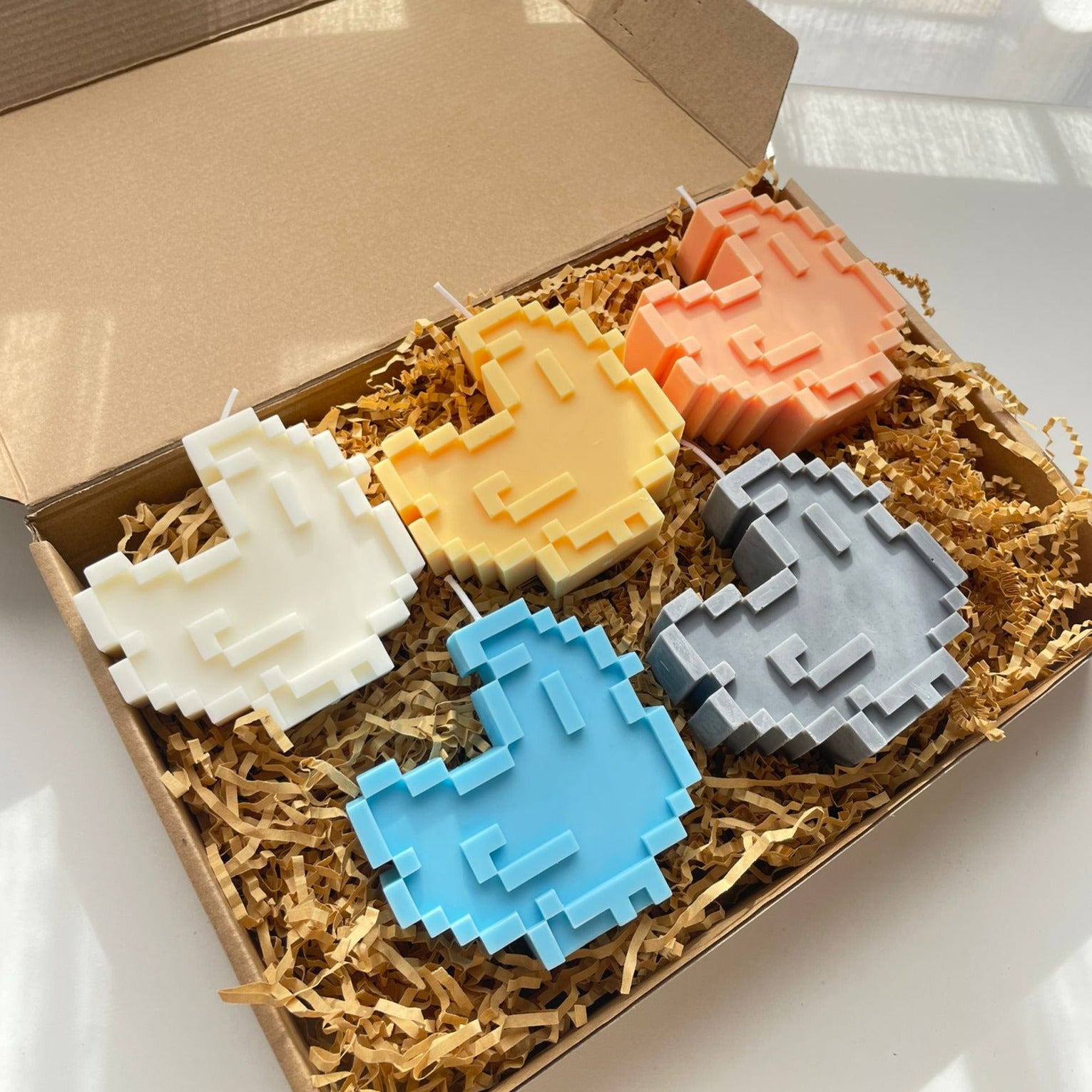 A cardboard box filled with shredded packing material contains six petimint Chicken Stardew Candles, each shaped like pixelated thumbs-up symbols. The candles come in white, orange, yellow, blue, gray, and light blue. The open box bathes in sunlight streaming in, perfect for adding a touch of gamer flair to your space.