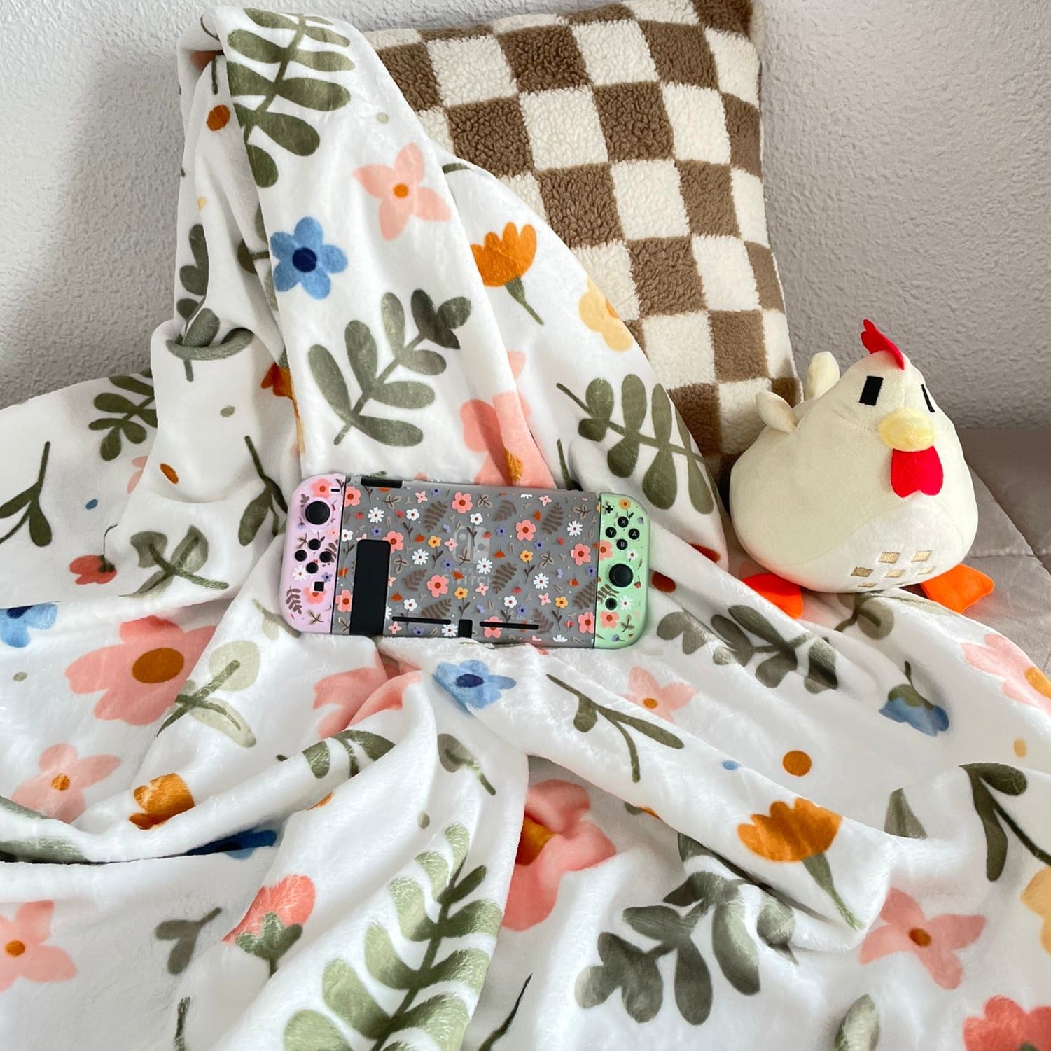 A cozygamer setup features the petimint Spring Blanket, a white blanket adorned with colorful floral and leaf patterns, a checkered brown and white pillow, a small plush chicken toy, and a Nintendo Switch with a floral case and pastel Joy-Cons in green and pink.