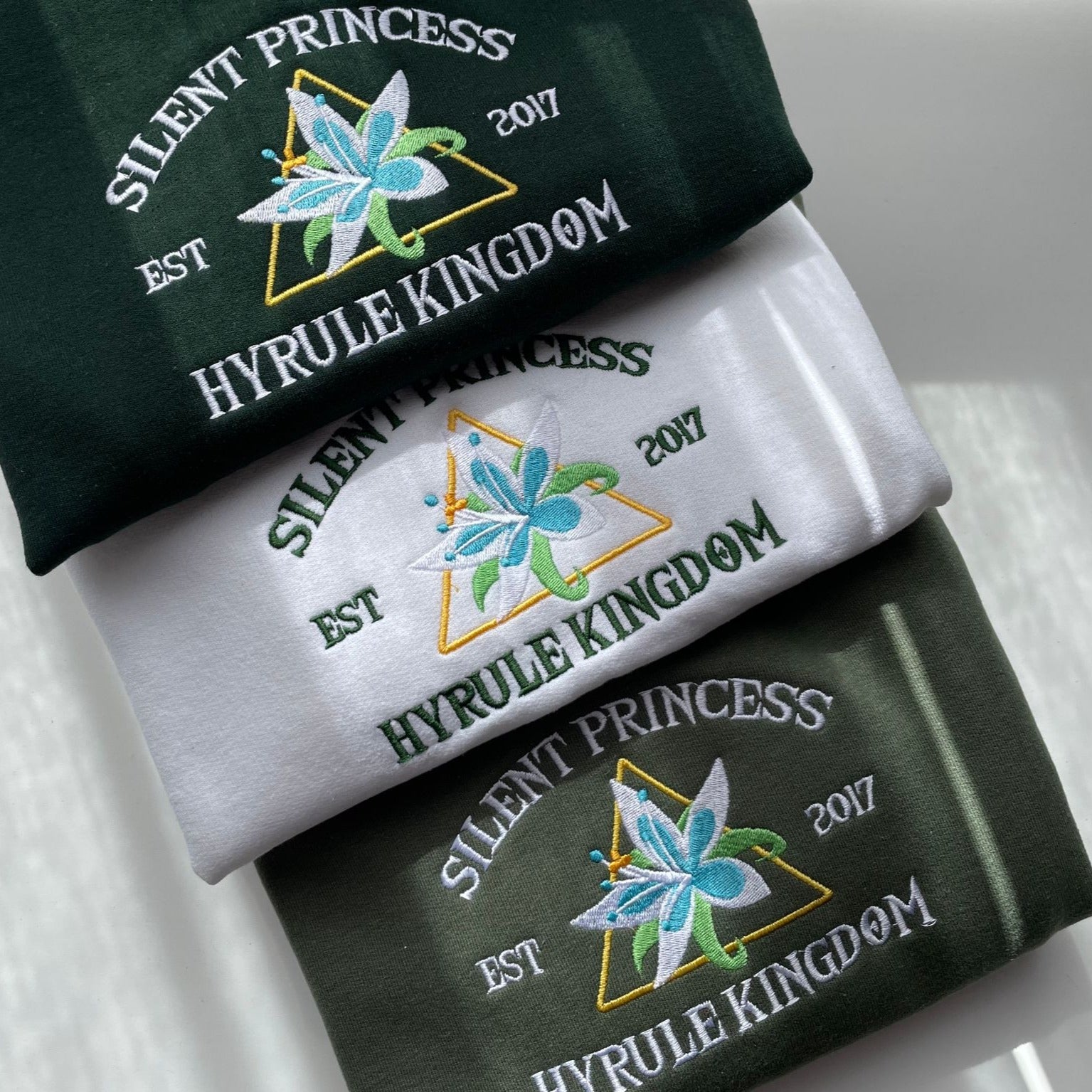 Three folded pieces of petimint's Silent Princess Crewneck Gamer Apparel in dark green, white, and dark green feature intricate embroidery with a blue floral design, a golden triangle, and the phrases "SILENT PRINCESS," "Hyrule Kingdom," and "EST 2017".