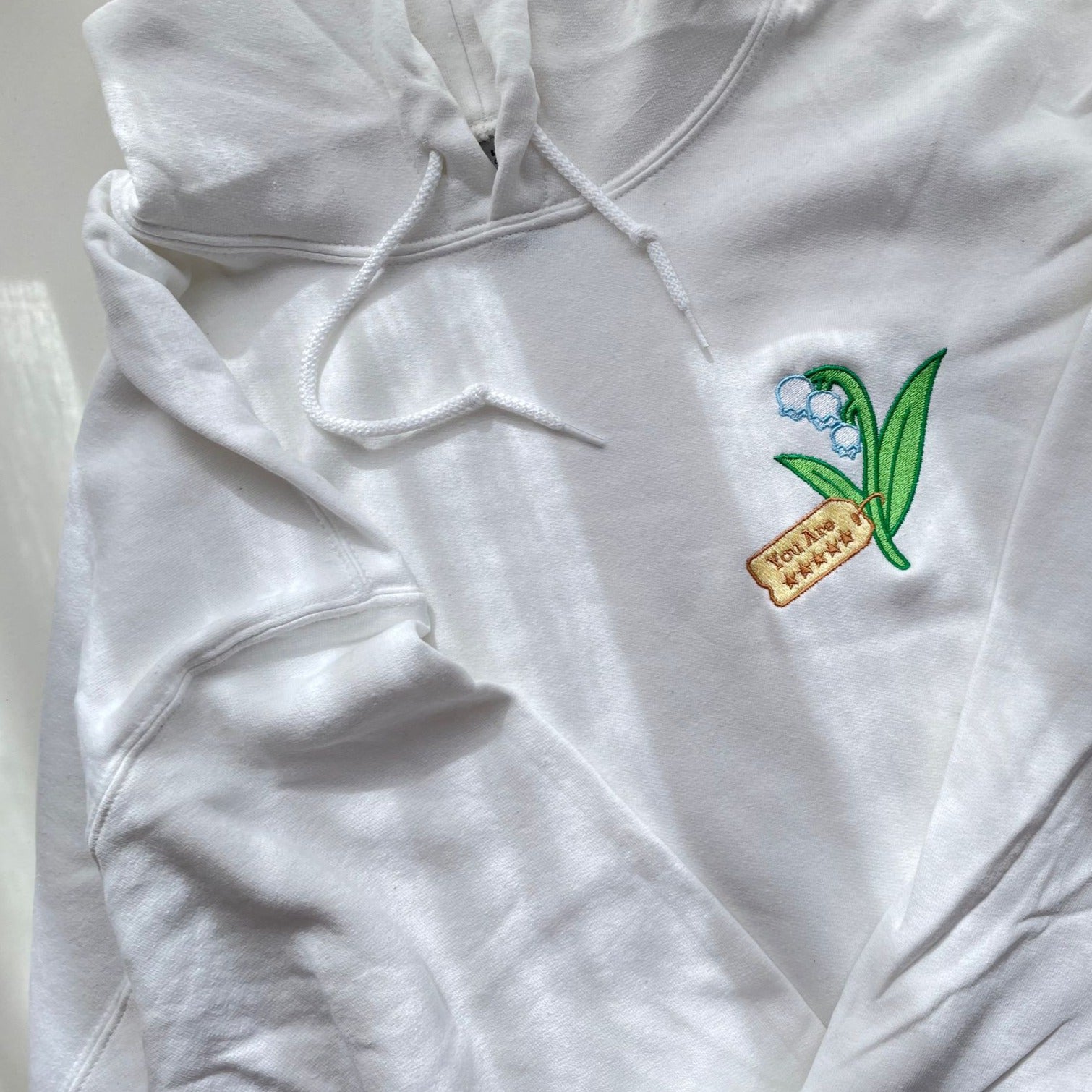 The Five Stars Hoodie by petimint is a white hoodie adorned with a small embroidered design on the chest, featuring a green lily of the valley plant and a brown tag with the word "Yoru" on it. This cozygamer essential has a drawstring hood and is displayed flat for your viewing pleasure.