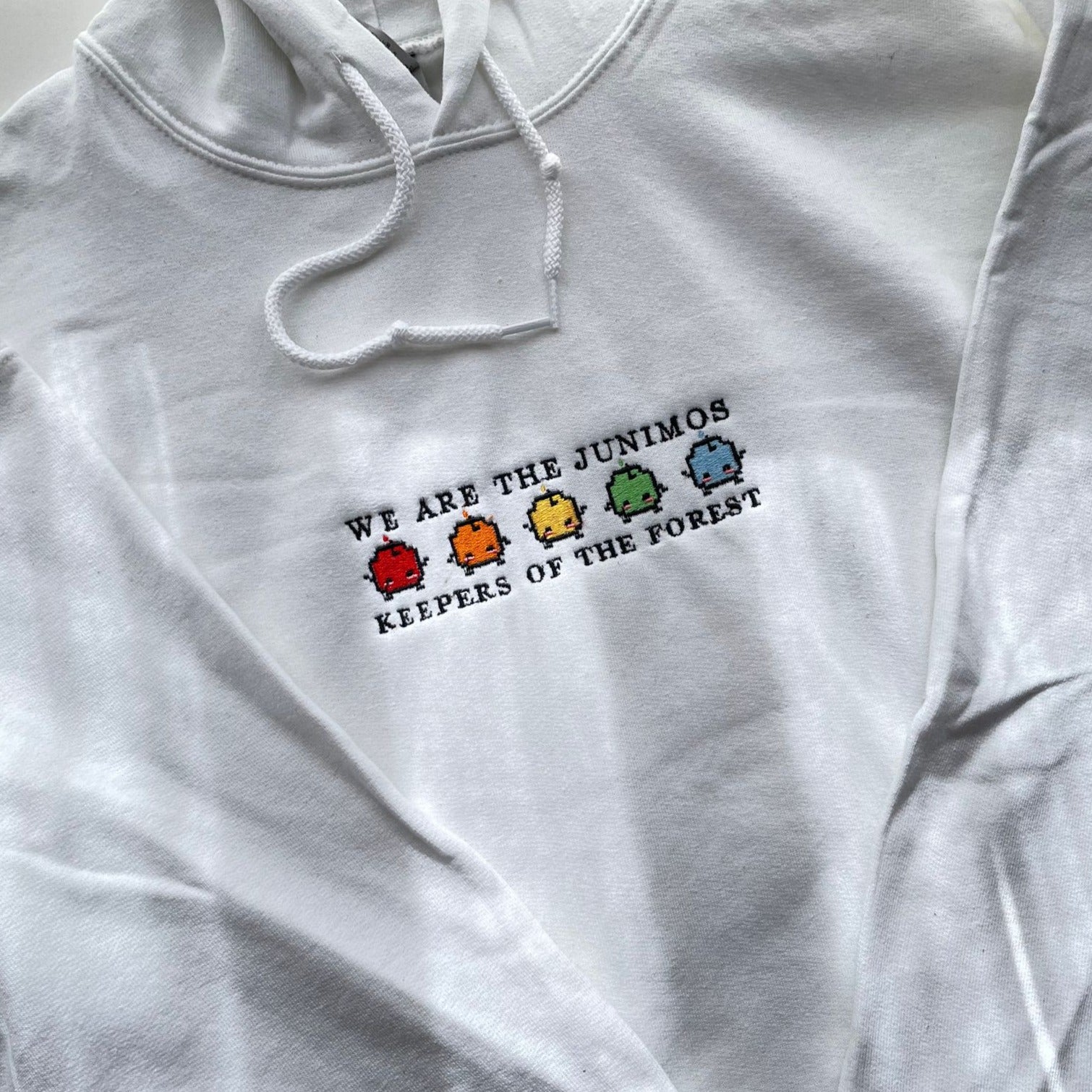 The Junimos Hoodie by petimint is a cozygamer apparel piece that features a white hoodie adorned with embroidered text and vibrant characters. The text reads "WE ARE THE JUNIMOS KEEPERS OF THE FOREST," alongside five small, pixelated, multicolored creatures arranged in a row.