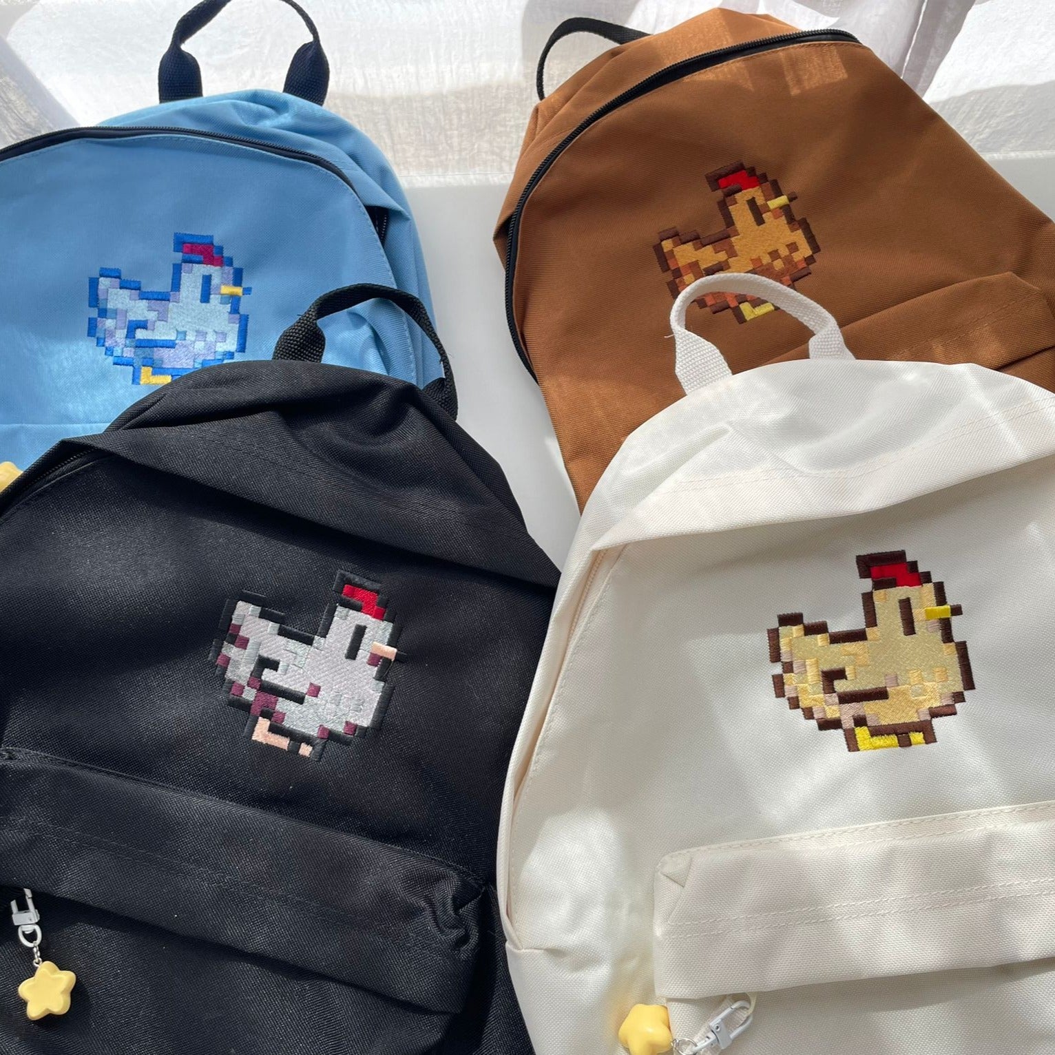 Four Stardew Chicken Backpacks from petimint are displayed on a surface, each featuring a pixelated chicken design. Perfect for the cozy gamer or gamer on the go, these backpacks come in blue with a blue chicken, brown with a yellow chicken, black with a white chicken, and white with a yellow chicken.