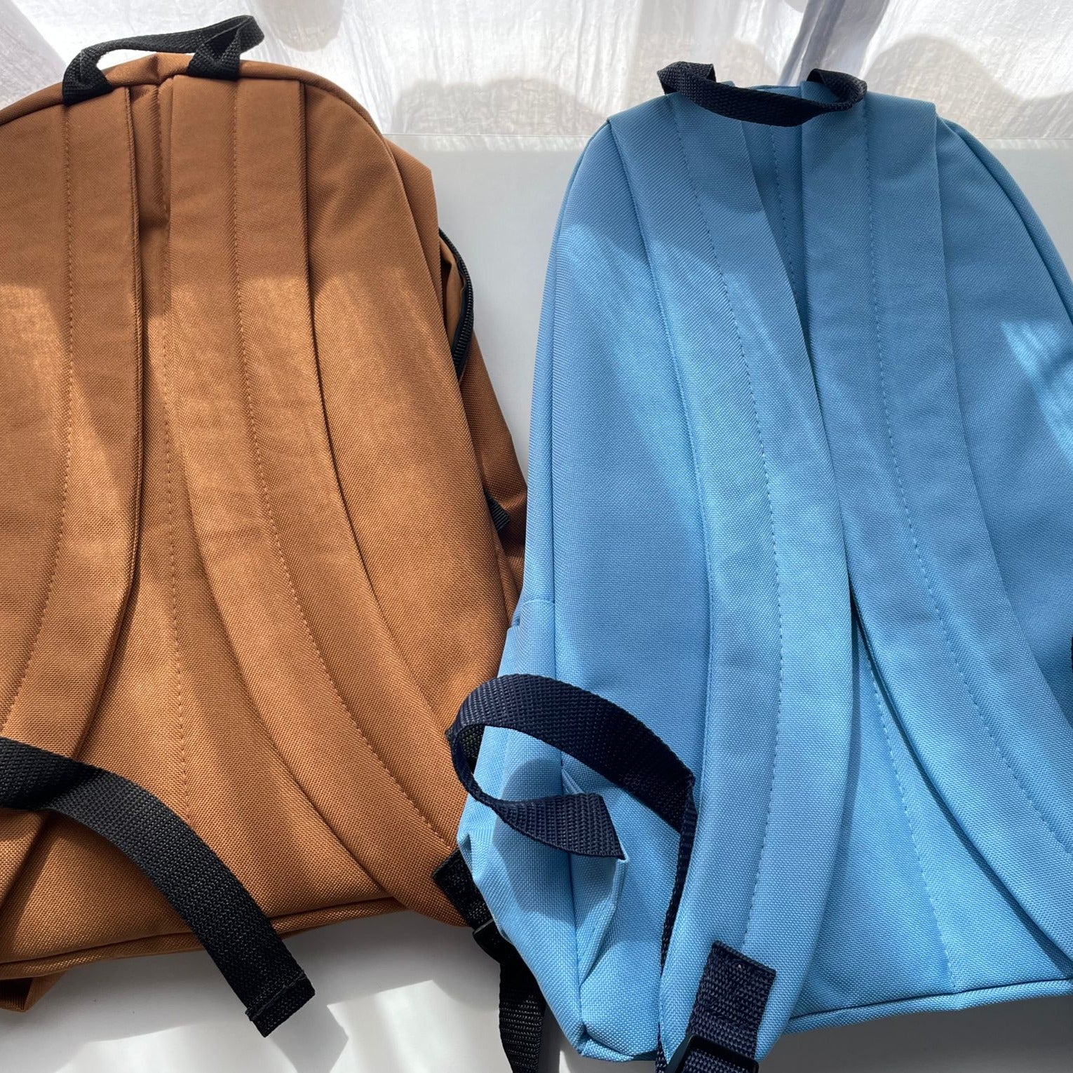 Two petimint Stardew Chicken Backpacks are pictured on a white surface, illuminated by natural light. The backpack on the left is brown with black straps, while the one on the right is light blue with black straps. Both have a quilted texture on their backs, making them perfect for carrying your cozygamer apparel.