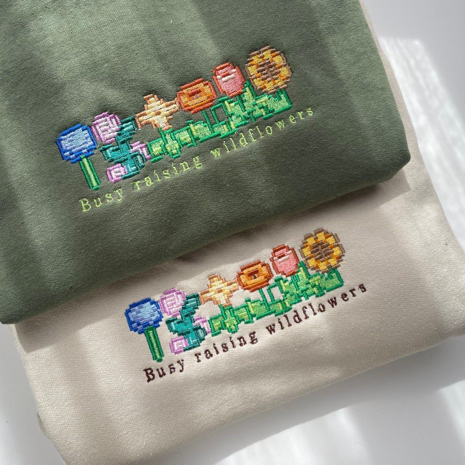 The Wildflowers Crewneck Gamer Apparel by petimint comes in two variations, one green and one beige, both showcasing vibrant embroidered flowers and the phrase "Busy raising wildflowers." This gaming apparel highlights various flowers in pink, red, yellow, blue, and a sunflower-like bloom.