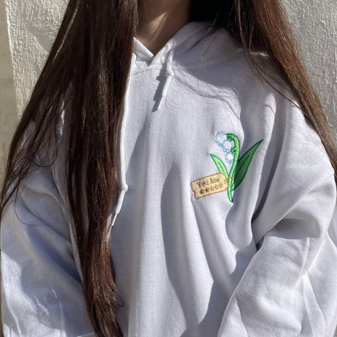 A person with long brown hair is wearing the Five Stars Hoodie by petimint, which features an embroidered design of a green plant with white flowers and a small beige tag with text. The background is a textured, off-white wall.