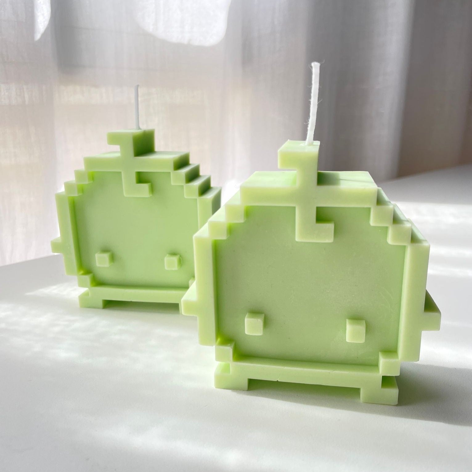 Two green, pixelated, 3D-printed Junimo Candles from petimint shaped like characters from a video game. Each candle features a face with two square eyes and a mouth. They stand on a light surface with soft light filtering through a sheer curtain in the background—a perfect touch for any gamer's decor.