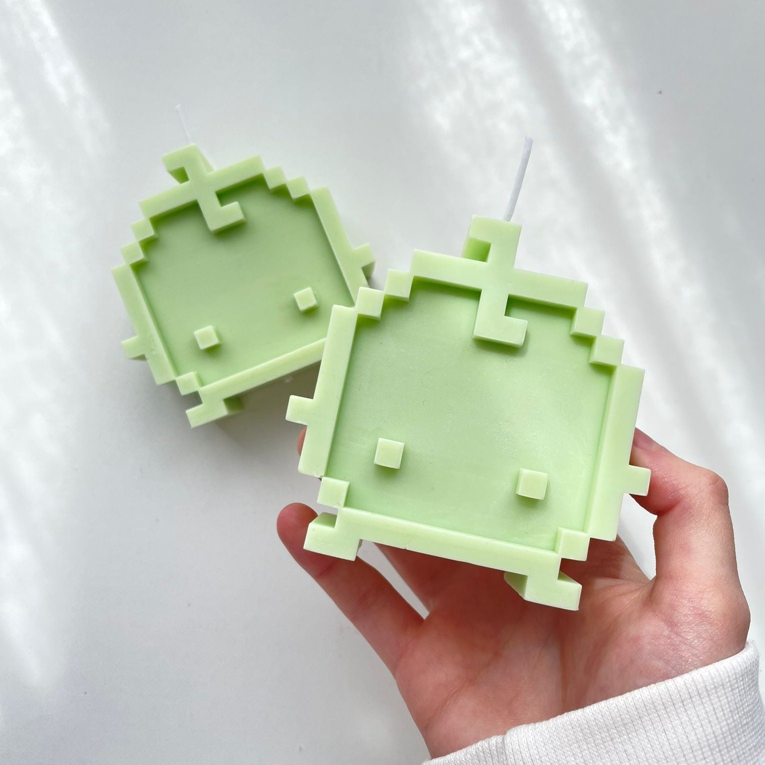 A hand holding one of two green, pixel art-style Junimo Candles from petimint against a white background. Perfect for the cozygamer, these candles feature square and rectangular shapes, enhancing their pixelated appearance.