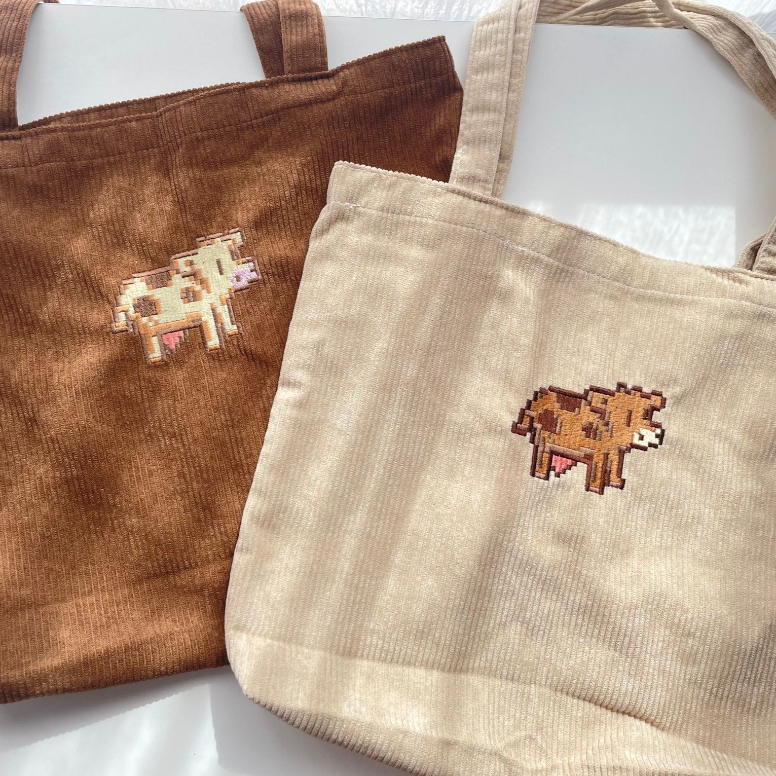 Two Stardew Cows Tote Bags by petimint, one in brown and the other in beige, are lying flat. Each tote features an embroidered pixel art-style cow in the center. The bags have wide handles and a textured corduroy fabric, ideal for your cozygamer apparel collection. The background is a white surface with subtle shadows.