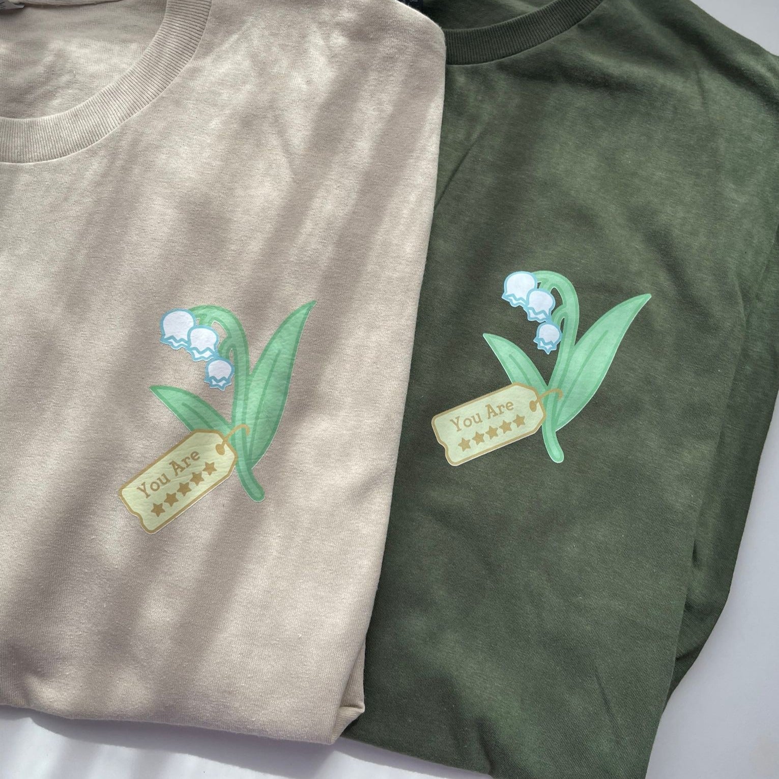 Two Five Stars T-Shirts by petimint are laid flat, one in beige and the other in olive green. Both pieces feature an embroidered design of lily of the valley flowers with a small sign that reads "You Are ★★★★★" near the stems and leaves. Sunlight casts soft shadows on the fabric, making them perfect for any cozy gamer.