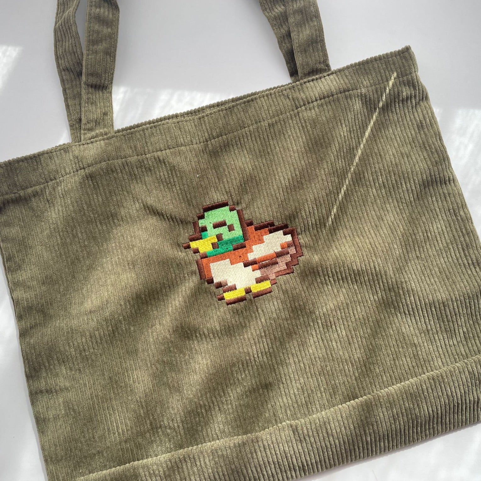 The Stardew Duck Tote Bag by petimint, crafted from khaki green corduroy fabric, is displayed on a flat surface. The front of the bag showcases a vibrant pixel art illustration of a duck, blending bright green, brown, white, and yellow hues. Perfect for the cozygamer, its detailed stitches and texture are clearly visible—a stylish addition for any Nintendo Switch enthusiast.