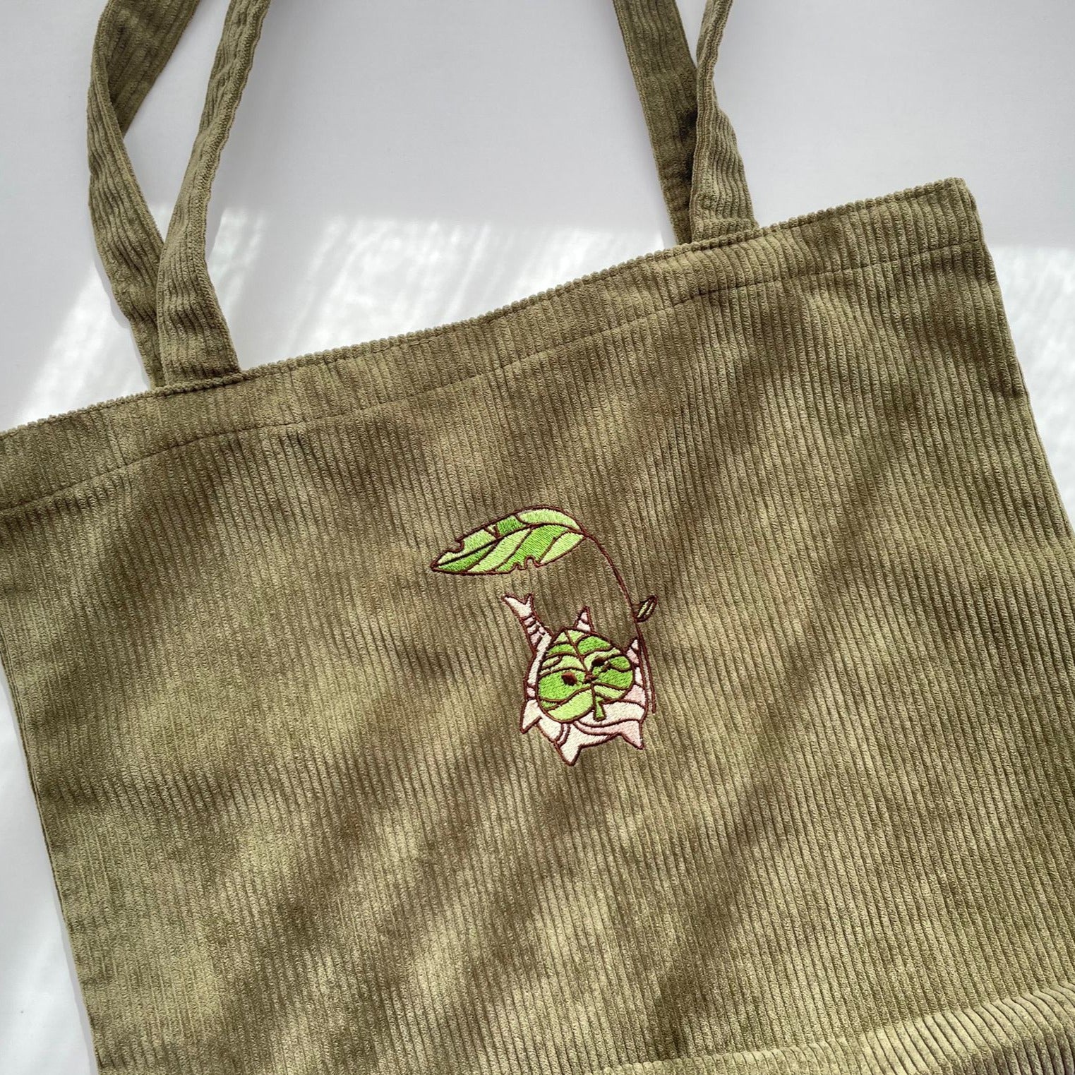 The Korok Tote Bag by petimint is a green corduroy tote bag with two straps and an embroidered design featuring a small, green, leaf-like character hanging upside down from a leafy branch. The whimsical, cartoonish character adds a cozygamer vibe to the plain fabric of the bag, making it perfect for any gaming enthusiast.