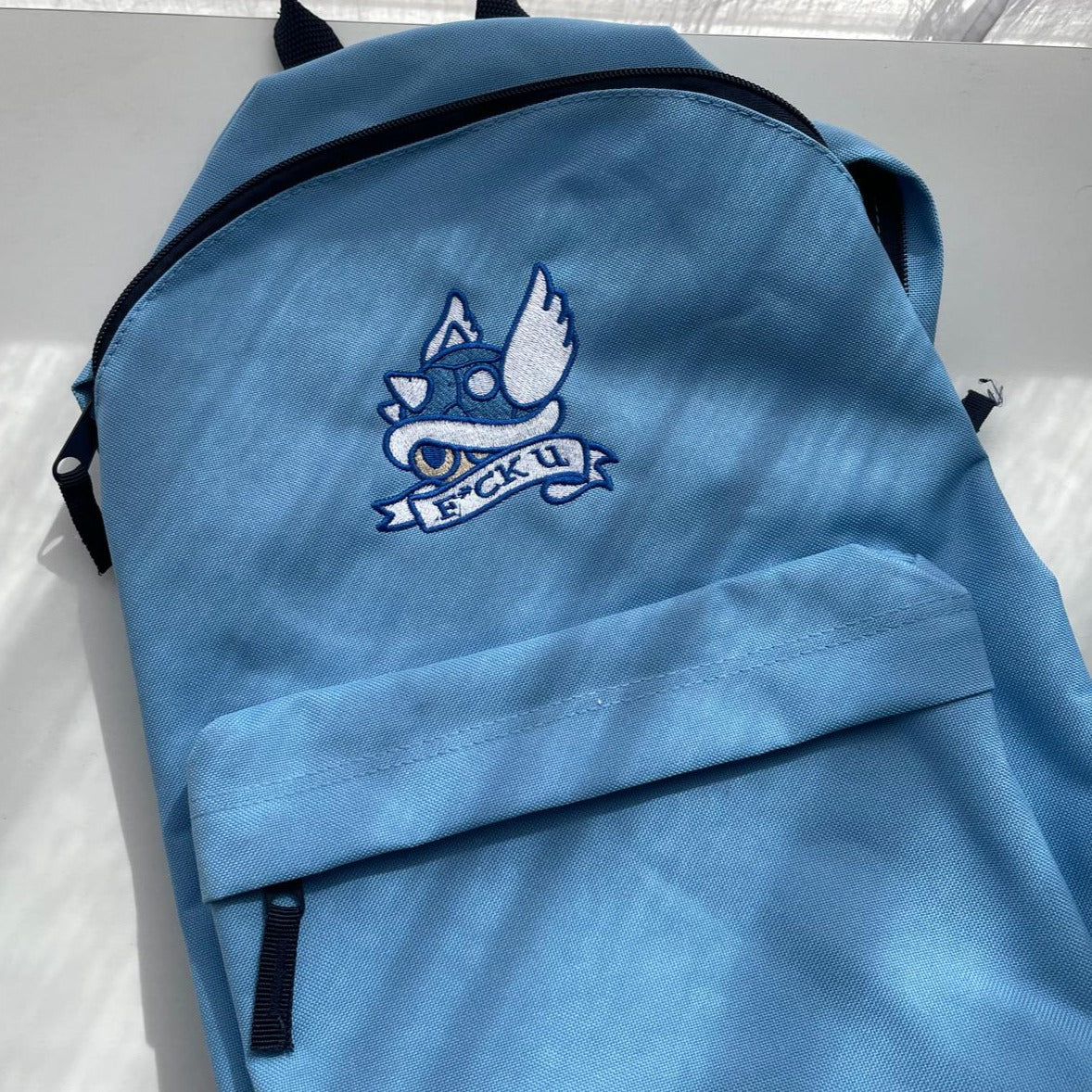 The petimint F*ck U Backpack Cozy Gamer Accessories features a light blue color, equipped with a front zippered pocket and adjustable shoulder straps. An embroidered patch depicting a blue shell with wings and sunglasses is prominently sewn onto the backpack, accompanied by a banner saying "FCK U," making it an ideal choice for the cozygamer on the move.