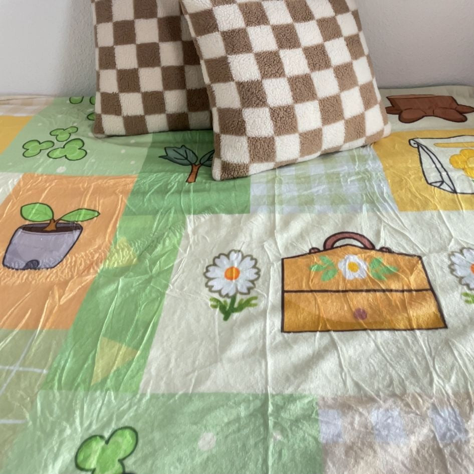 A cozy bed is adorned with the Cottagecore ACNH Blanket by petimint, a colorful quilt featuring various designs such as daisies, leaves, and potted plants. Two checkered brown and white pillows are propped up against the white wall at the head of the bed. The room's home decor creates a light and inviting atmosphere.