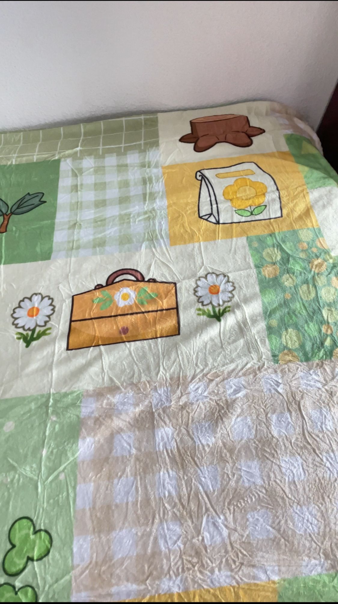The Cottagecore ACNH Blanket by petimint is draped invitingly on the bed, ideal for infusing a quaint charm into any home decor. This cozy quilt showcases an array of patterns featuring flowers, a suitcase, hat, and a bag adorned with a yellow flower. The color palette includes soothing shades of green, yellow, and beige interspersed with checkered and solid blocks.