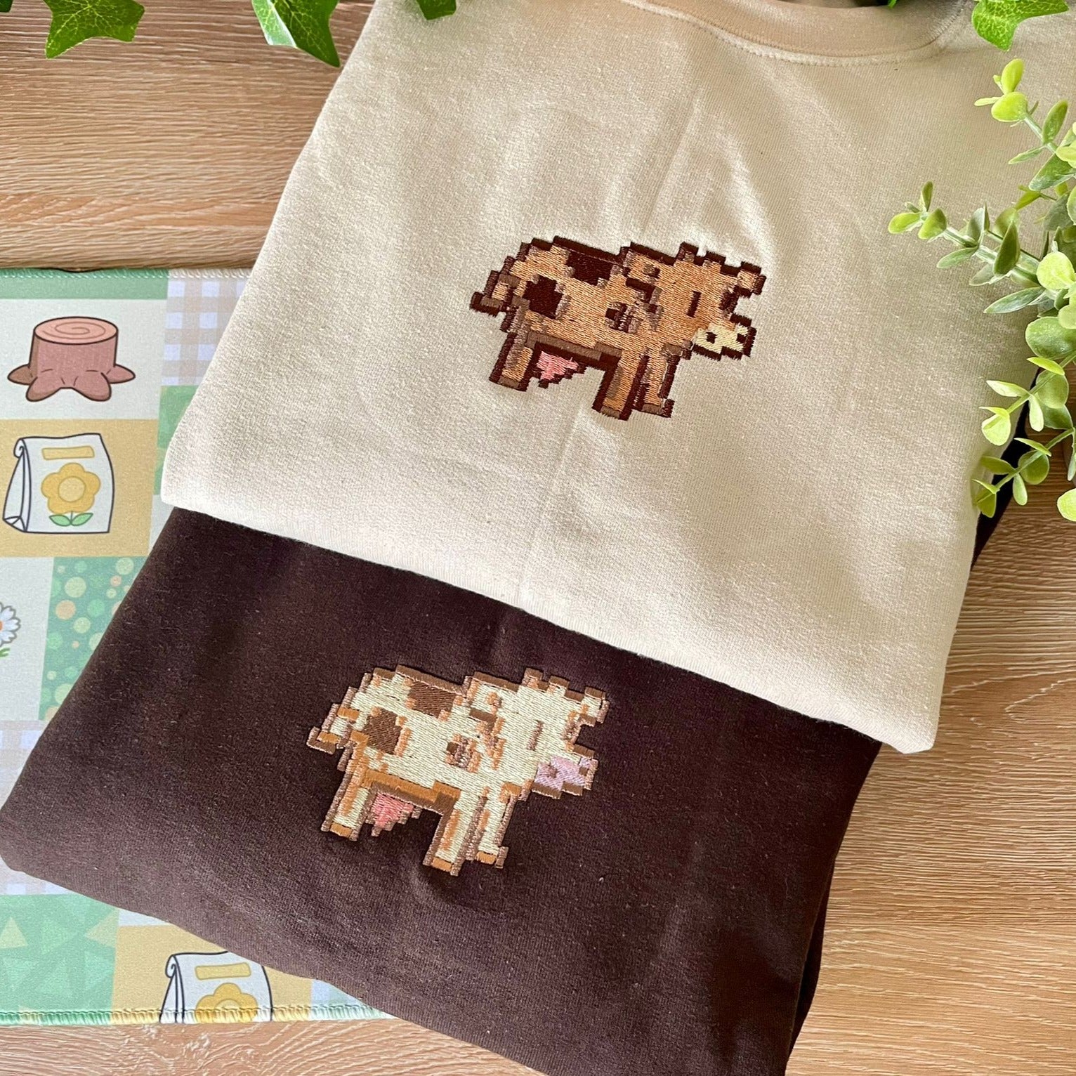 Two folded "Stardew Cow Crewneck" sweatshirts from petimint are displayed on a wooden surface, one in beige and the other in dark brown, each featuring a pixelated cow graphic on the front. Greenery is thoughtfully arranged around the apparel, and a patterned cloth adorned with tree stumps and honey jars is partially visible. Perfect for bringing out the cozygamer in you.