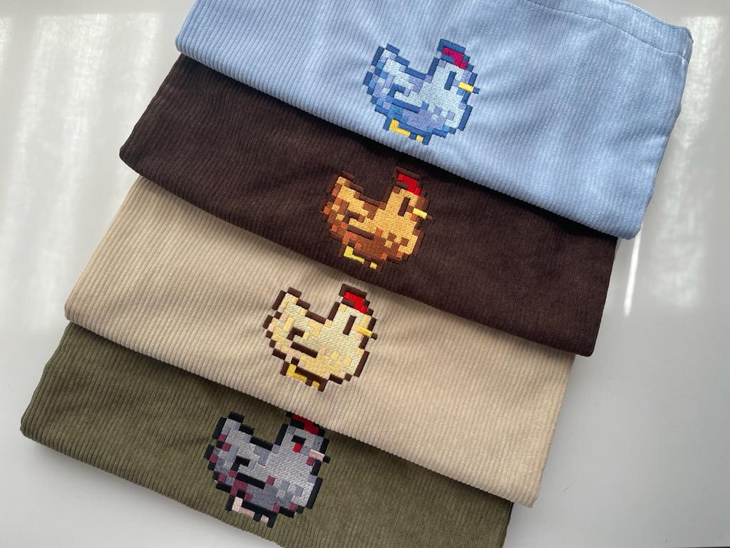 Four neatly folded corduroy fabrics with embroidered pixel art chickens, reminiscent of charming gaming aesthetics. The fabrics are stacked in a gamer-friendly palette: light blue, dark brown, beige, and olive green. Each fabric showcases unique chicken designs perfect for stylish apparel.