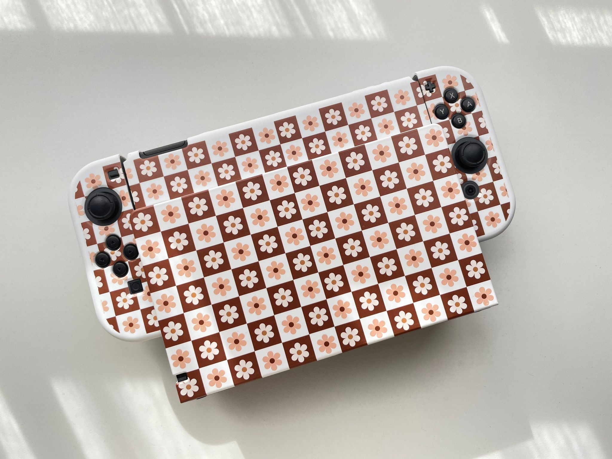 A handheld gaming console with a retro floral and checkerboard pattern skin. The pattern consists of alternating brown and white squares, each adorned with a small white flower. Perfect for the cozy gamer, the console boasts black analog sticks and buttons, merging style with comfort.