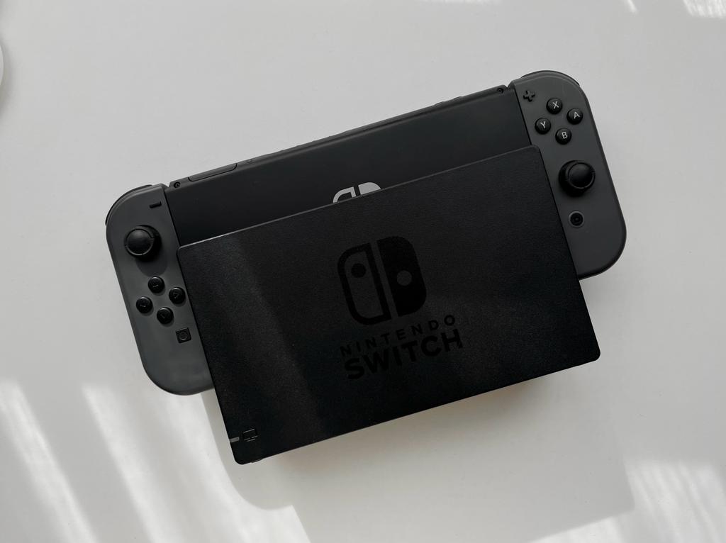 A Nintendo Switch console in docked mode is shown from a slightly high angle. The screen is partially obscured by the dock, displaying the Nintendo Switch logo. The Joy-Con controllers are attached on both sides, and everything is resting on a white surface. It’s an ideal setup for a cozy gamer.