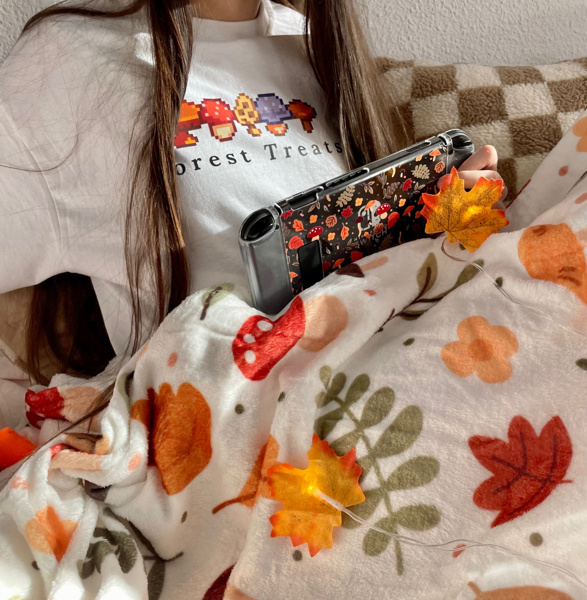 A person with long brown hair is wearing a white shirt with "Forest Treats" and mushroom graphics on it. Covered in a blanket adorned with various autumn-themed patterns, this cozygamer is engrossed in playing on their Nintendo Switch, which also features a mushroom-patterned cover.