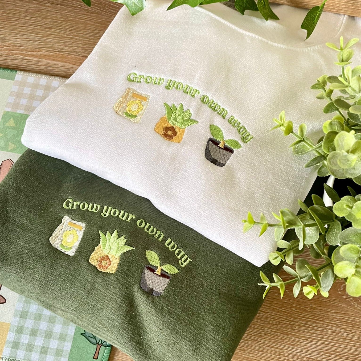 Two Grow ACNH Crewneck sweaters from petimint, one in white and one in green, both featuring embroidered designs of plants and the words "Grow your own way." Laid out on a table near leafy plants, these cozygamer sweaters are perfect for gaming sessions. Each design includes a plant in a jar, a potted plant, and a small plant.
