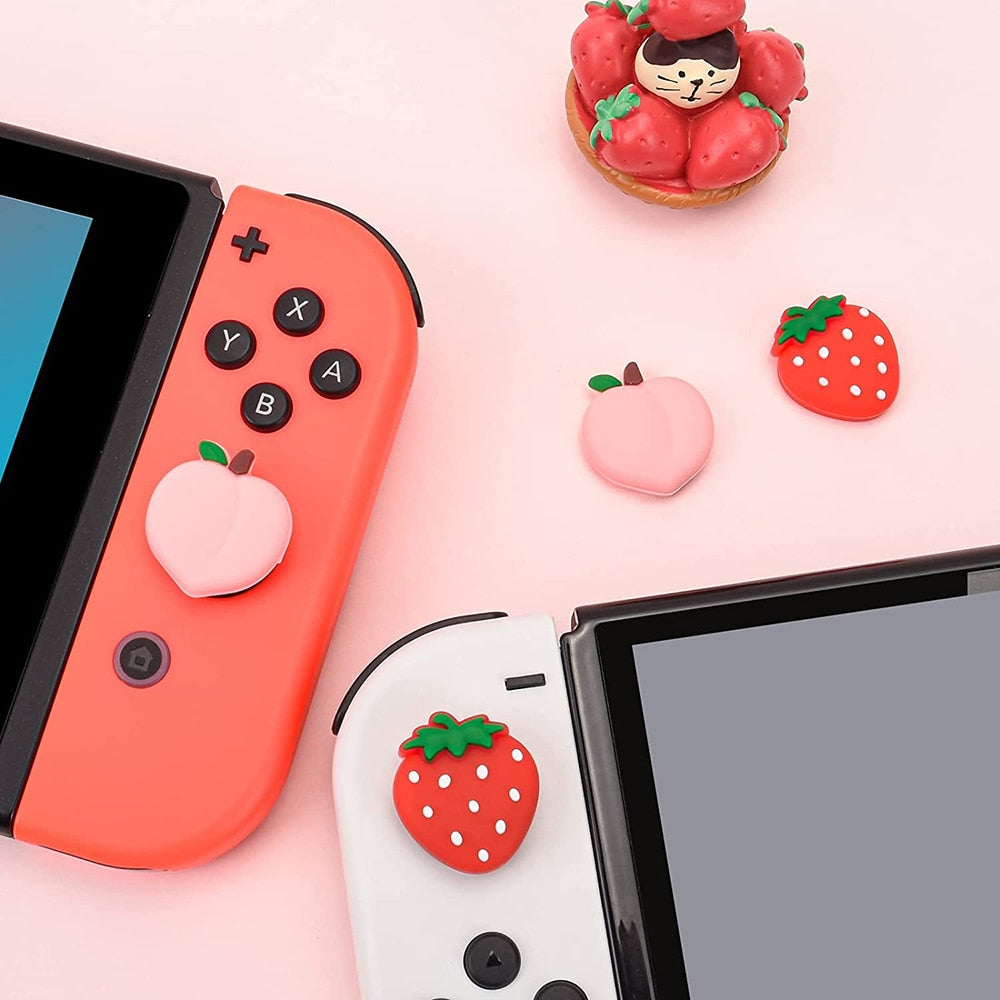 Two gaming consoles, similar to a Nintendo Switch, feature detachable controllers adorned with petimint's Strawberry and Peach Thumb Grips: a peach on the red controller and a strawberry on the white controller. Nearby on a pink surface lie additional strawberry and peach thumb grips from petimint, along with a small cat figurine surrounded by strawberries—a setup any gamer would adore.