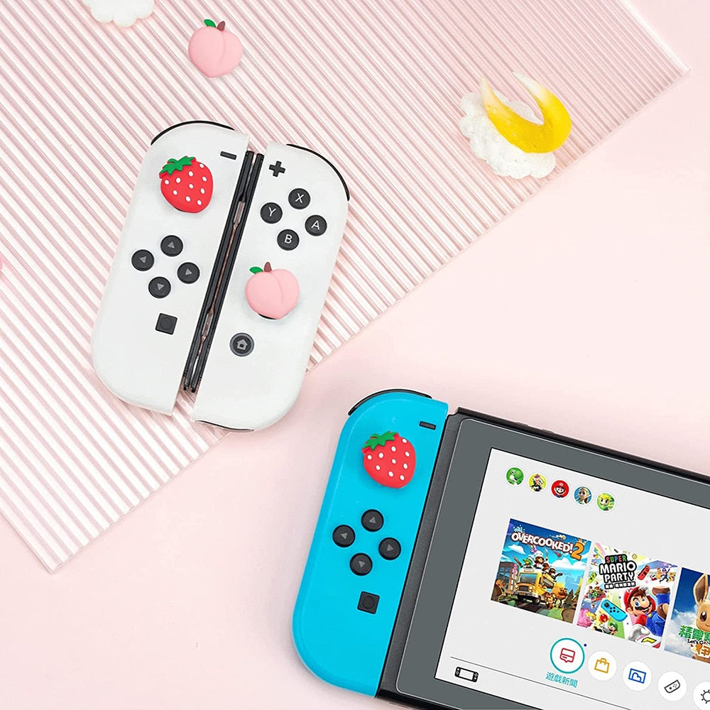 A Nintendo Switch console featuring petimint's Strawberry and Peach Thumb Grips, with a blue left Joy-Con adorned with a strawberry sticker and a white right Joy-Con embellished with strawberry and peach stickers. The screen displays the game library, perfect for any cozy gamer. The Joy-Cons rest on a striped surface accompanied by additional fruit-themed decorations.