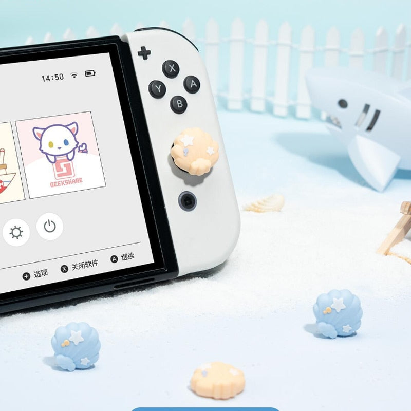 A Nintendo Switch, adorned with petimint's Seashell Thumb Grips, is presented on white sand. In the background, a toy shark and starfish enhance the scene alongside a white fence and blue sky. The console's screen showcases icons and app options in a playful setup.