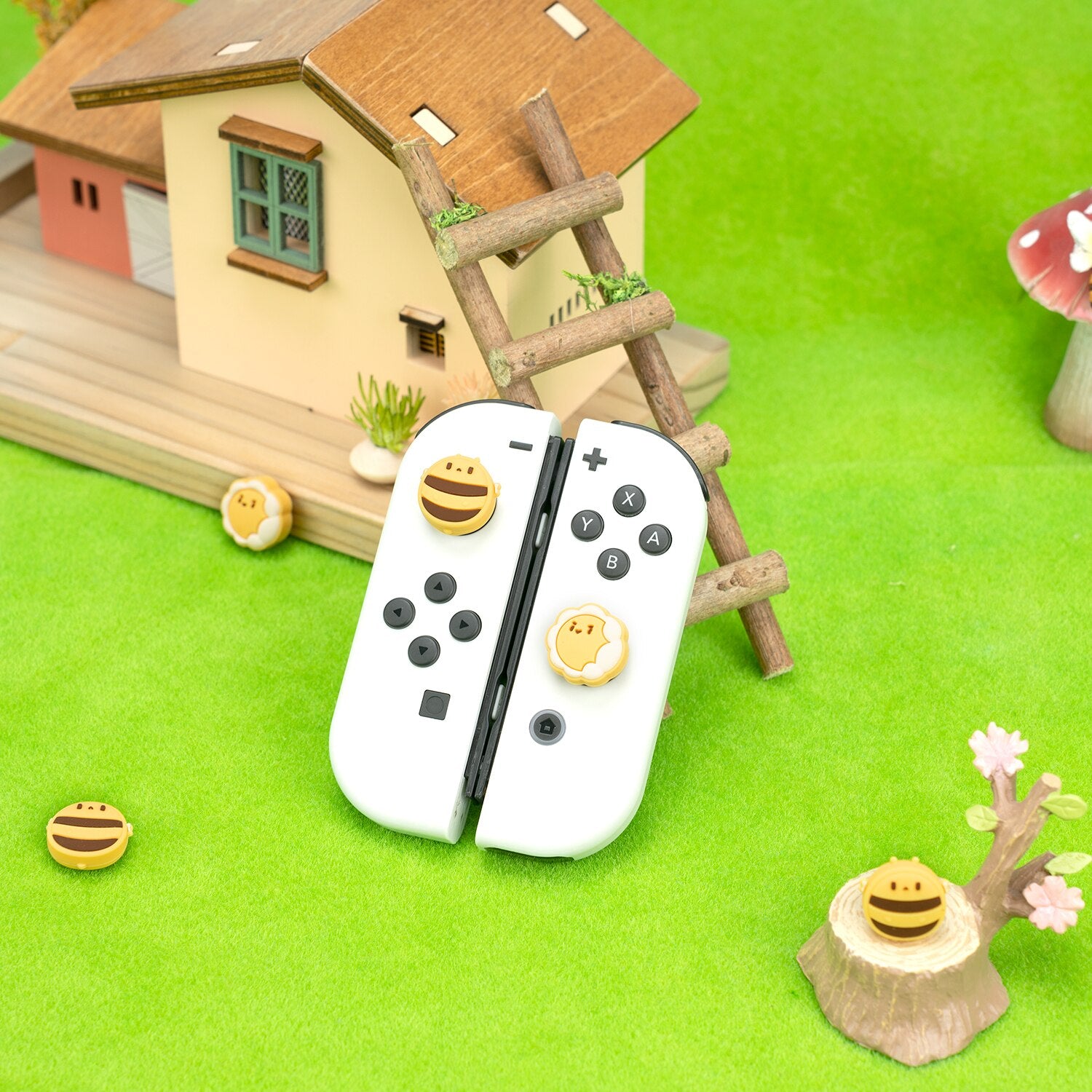 Petimint's Bees Thumb Grips for Nintendo Switch are beautifully showcased resting on white gaming controllers against a backdrop of green grass and a small model house featuring a wooden ladder. A large mushroom adds to the whimsical, playful scene, making it ideal for Nintendo Switch enthusiasts.