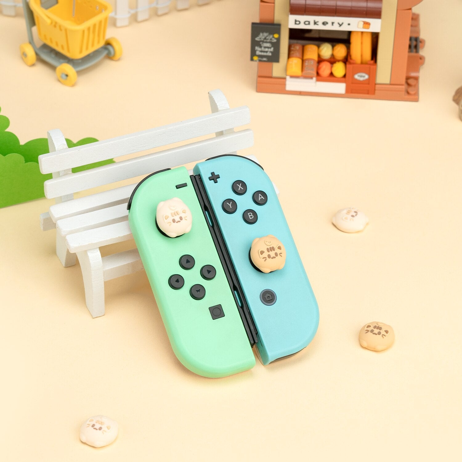 A pair of pastel-colored Soft Cats Thumb Grips for Nintendo Switch, one green and one blue, from the brand petimint, are placed on a miniature white bench. The thumb grips resemble small cookies with smiley faces on their buttons. Set against the backdrop of a toy bakery and cart, this scene exudes playful charm for any cozygamer.