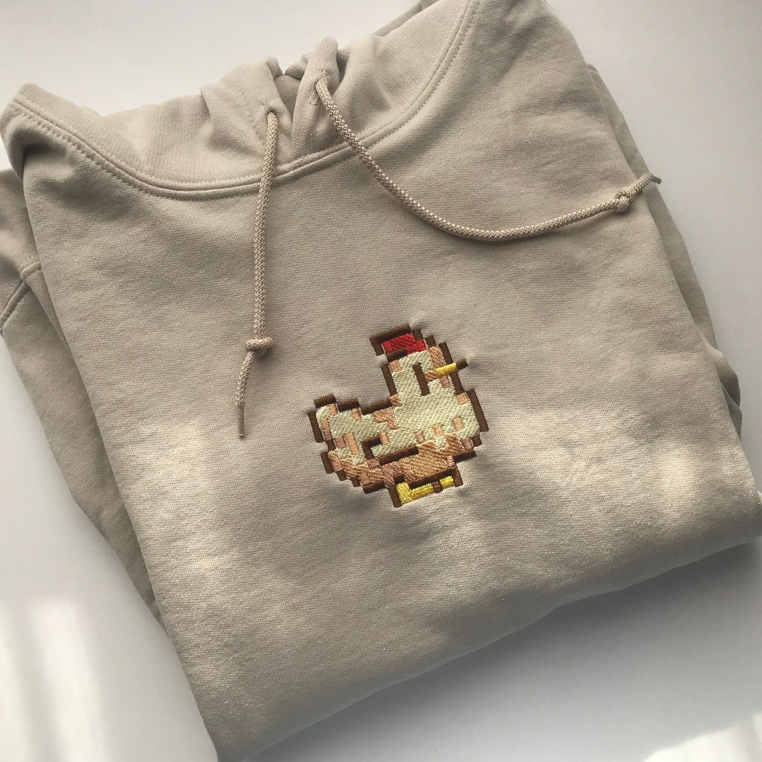 Discover the inviting allure of petimint's Stardew Chicken Hoodie Apparel, adorned with intricate embroidered pixel art showcasing a chicken in cozy tones of brown, red, yellow, and cream. This unisex beige hoodie is elegantly displayed on a sleek, light background.