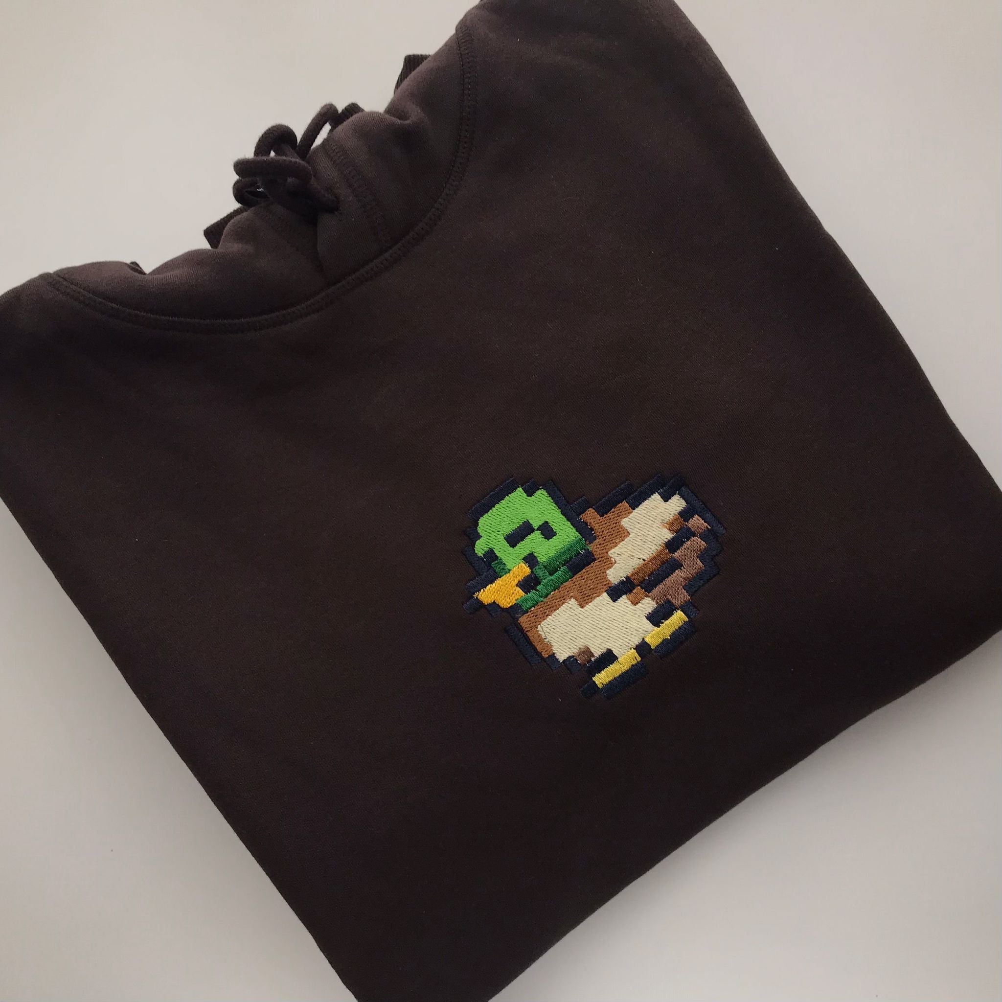 The Stardew Duck Hoodie Apparel by petimint is a stylish folded black sweatshirt, featuring a pixelated image of a duck on the chest with a green head, yellow beak, and brown and white body. Perfect for gaming enthusiasts, this piece of apparel is neatly arranged on a light grey surface.