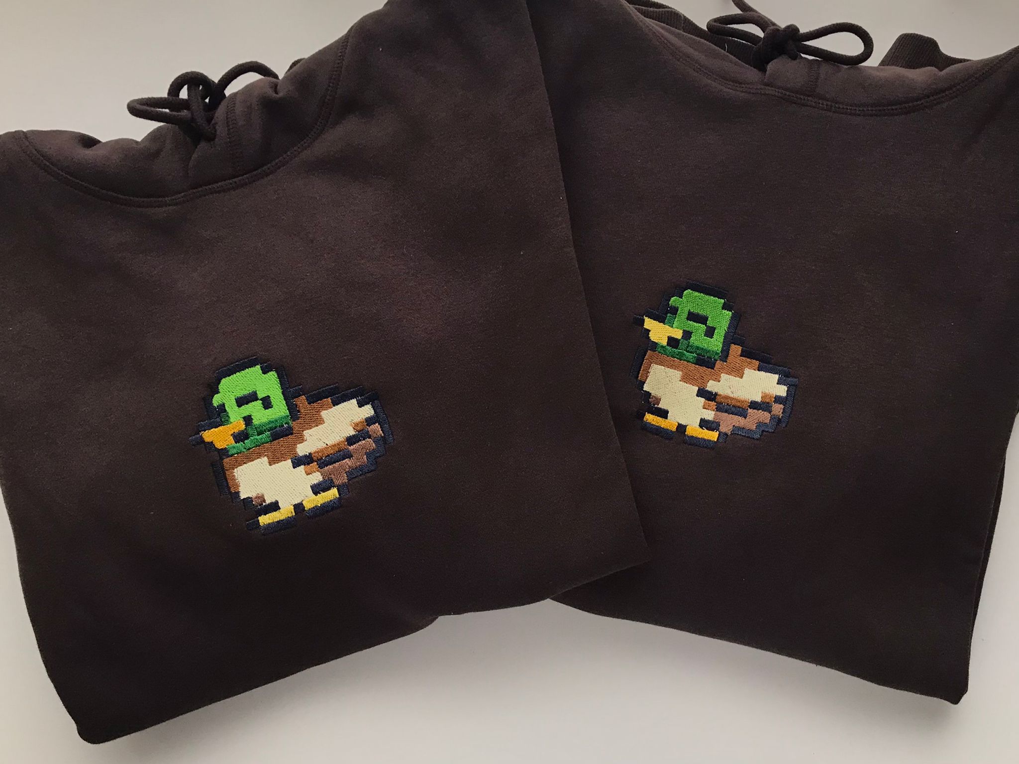 Two black Stardew Duck Hoodies by petimint lie flat side by side. Each hoodie features a cute embroidered design of a green-headed duck with a brown body on the chest. Perfect for the cozygamer, these gaming apparel essentials also showcase visible drawstrings at the top.
