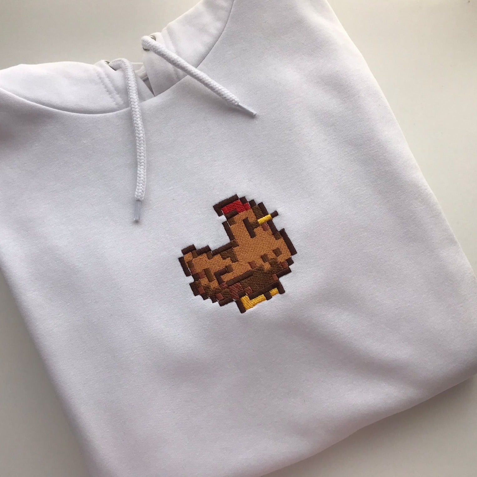 The Stardew Brown Chicken Hoodie by petimint showcases a neatly folded, white design featuring an embroidered, small pixelated brown chicken with a red comb and yellow feet on the chest. Perfect for any gamer, this stylish piece of apparel is completed with white drawstrings on the hood.