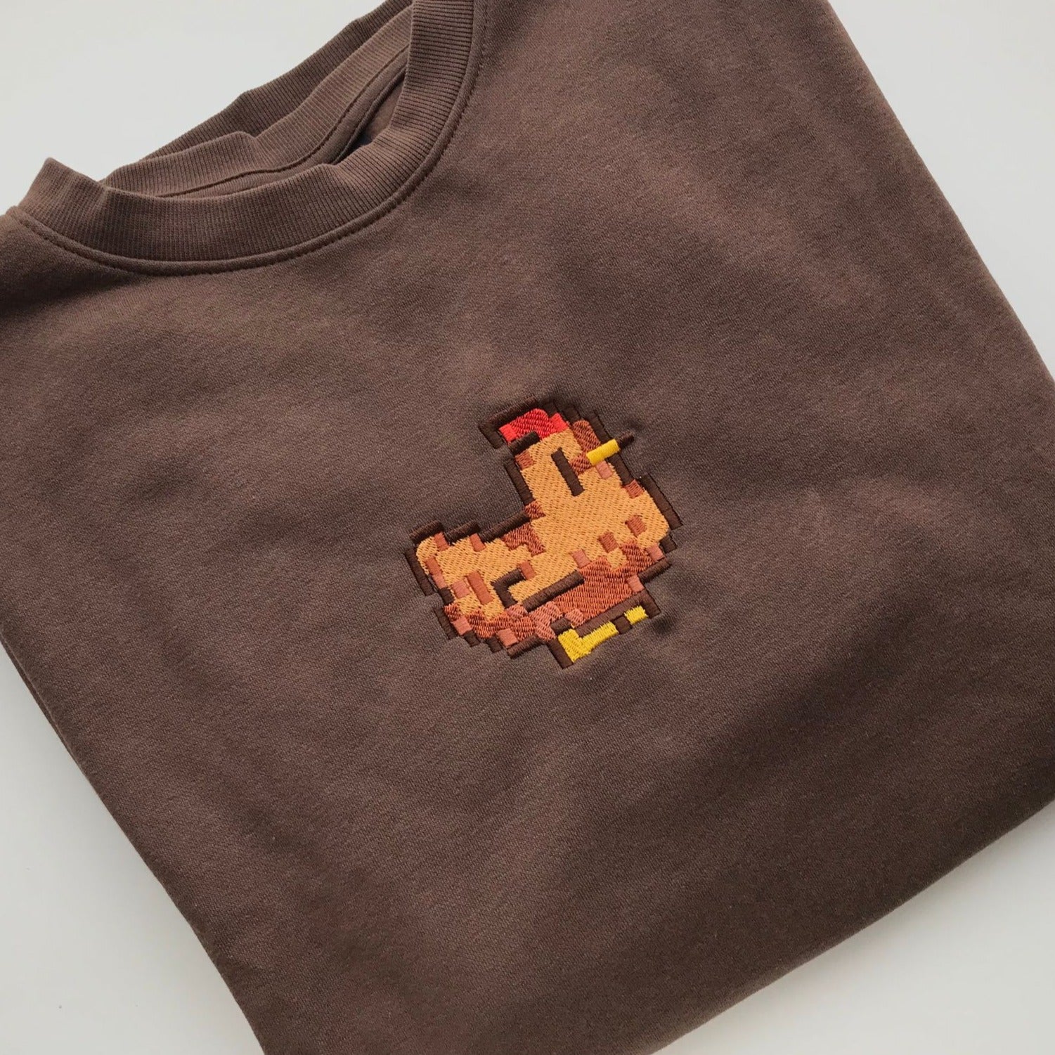 A neatly folded Stardew Brown Chicken Crewneck from petimint, featuring an embroidered pixel art chicken on the chest. The pixelated chicken, with its red comb, orange beak, and yellow feet, adds a playful and retro touch to the gaming-inspired apparel.